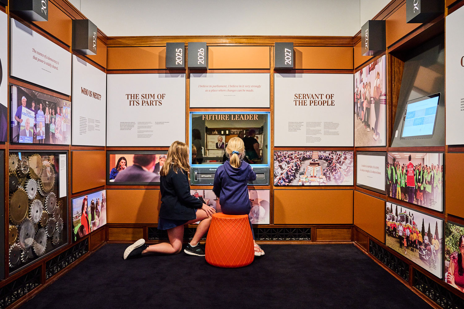 Two girls look into a mirror that says 'Future Leader' in an alcove suurrounded by exhibition panels with titles such as Servant of the people and dates like 2025, 2027.