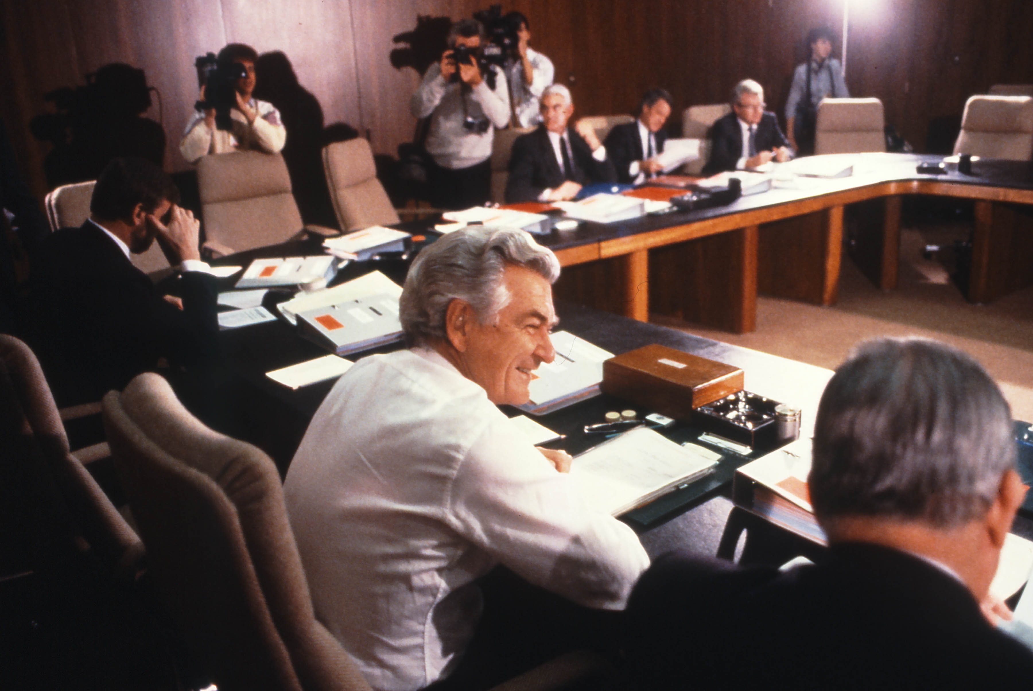This colour photograph is taken from behind Deputy Prime Minister Lionel Bowen who is seated beside Prime Minister Bob Hawke. Both are dressed in standard business attire. Other members of Cabinet can be seen sitting around the table although only half of the seats are filled. In the background are a photographer and two cameramen photographing and filming the cabinet meeting. This colour photograph is taken from behind Deputy Prime Minister Lionel Bowen who is seated beside Prime Minister Bob Hawke. Both are dressed in standard business attire. Other members of Cabinet can be seen sitting around the table although only half of the seats are filled. In the background are a photographer and two cameramen photographing and filming the cabinet meeting.