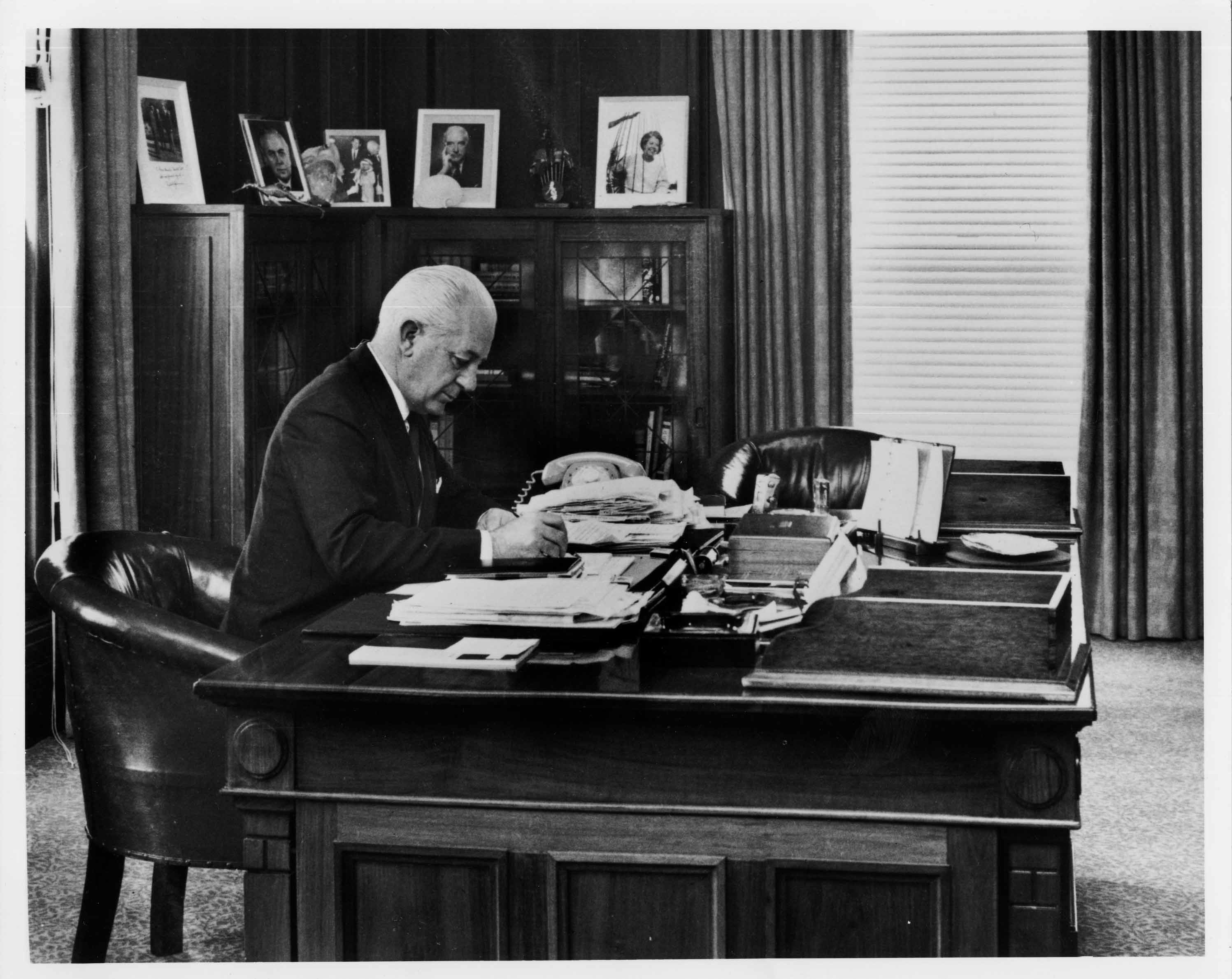 In this black and white photograph Prime Minister Harrold Holt sits at the timber desk made in 1926. Holt is wearing a dark suit, white shirt and tie and is leaning on the desk while he writes. The desk has fine carved panels with a geometric detailing on the corners. The surface of the desk is cluttered with stacks of documents and a telephone. On the glass-fronted cabinets in the background are a number of framed photographic portraits. In this black and white photograph Prime Minister Harrold Holt sits at the timber desk made in 1926. Holt is wearing a dark suit, white shirt and tie and is leaning on the desk while he writes. The desk has fine carved panels with a geometric detailing on the corners. The surface of the desk is cluttered with stacks of documents and a telephone. On the glass-fronted cabinets in the background are a number of framed photographic portraits.