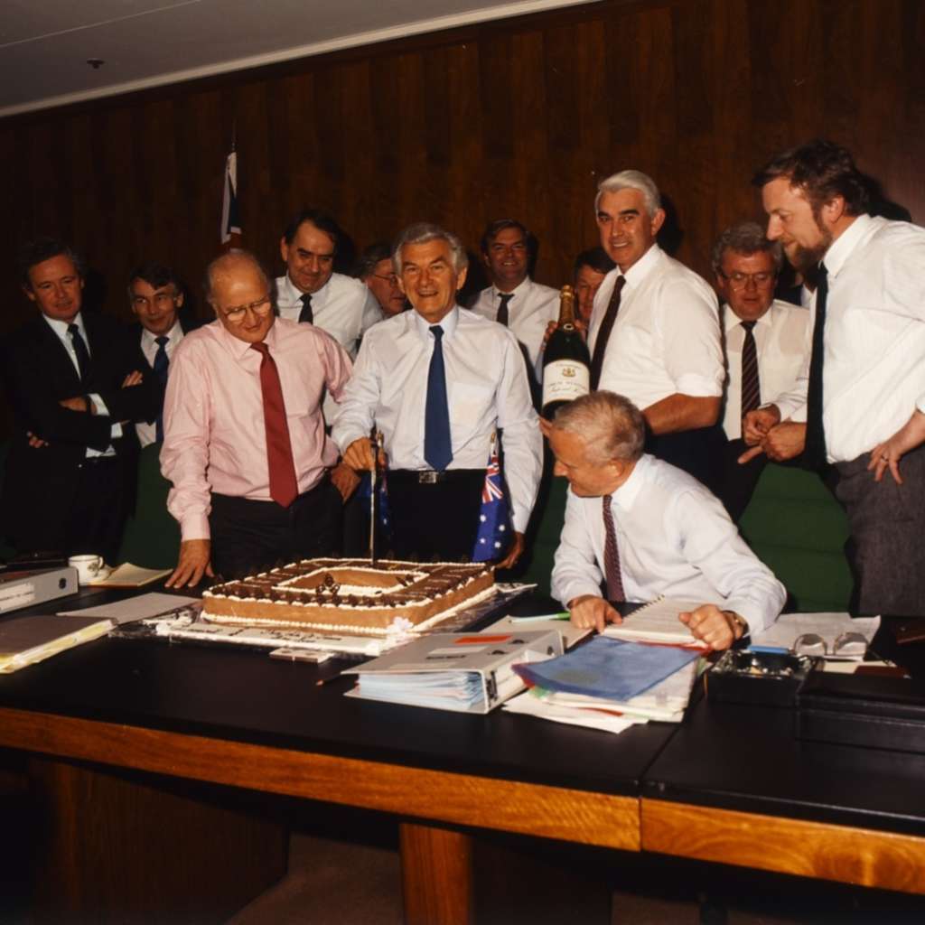 Prime Minister Bob Hawke stands and cuts a square cake, surrounded by a group of men all smiling. Prime Minister Bob Hawke stands and cuts a square cake, surrounded by a group of men all smiling.