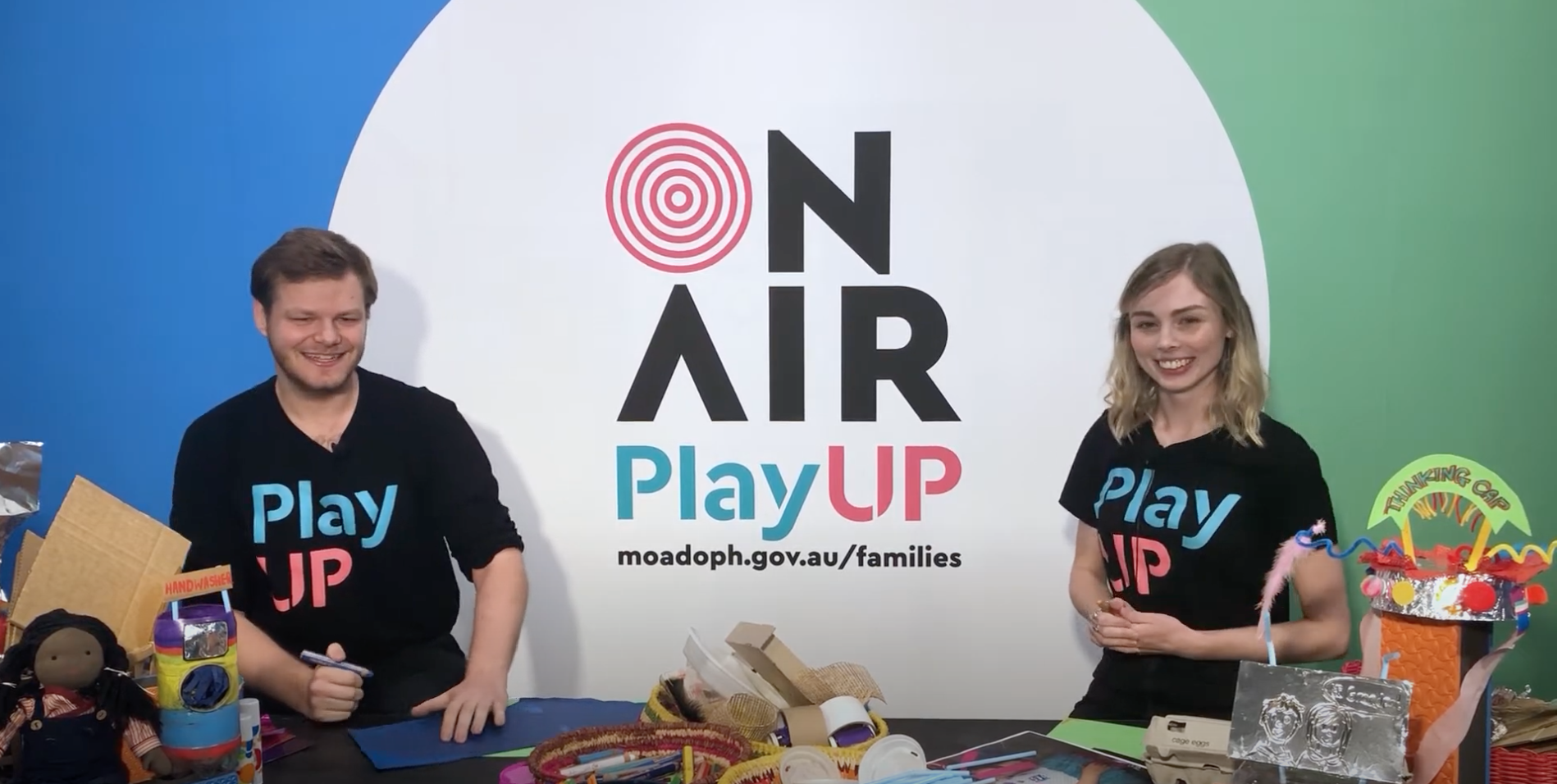 Two PlayUP facilitators stand behind a table covered in craft supplies. Two PlayUP facilitators stand behind a table covered in craft supplies.