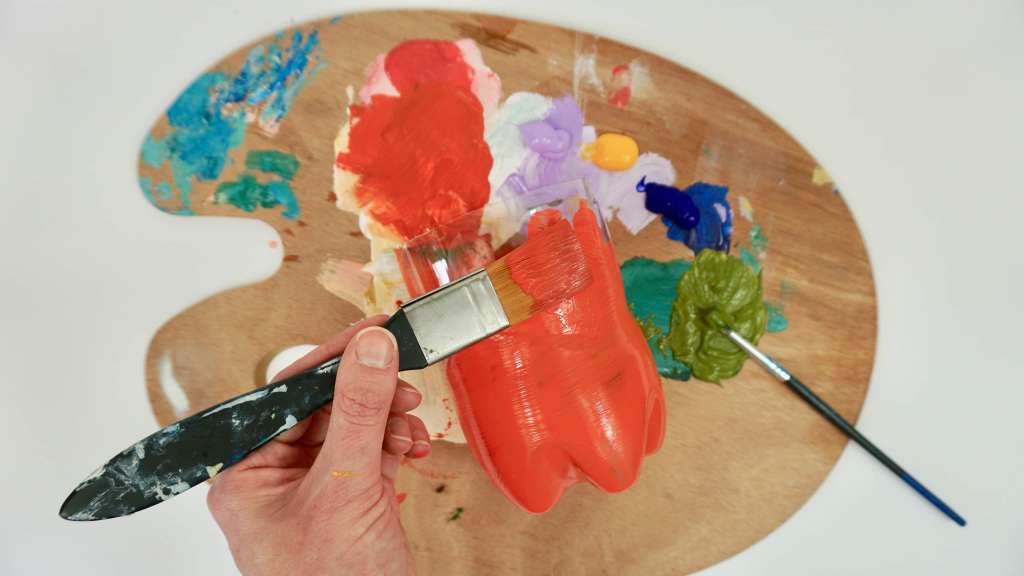 A close up of a hand holding a paint brush painting a plastic bottle red. A close up of a hand holding a paint brush painting a plastic bottle red.