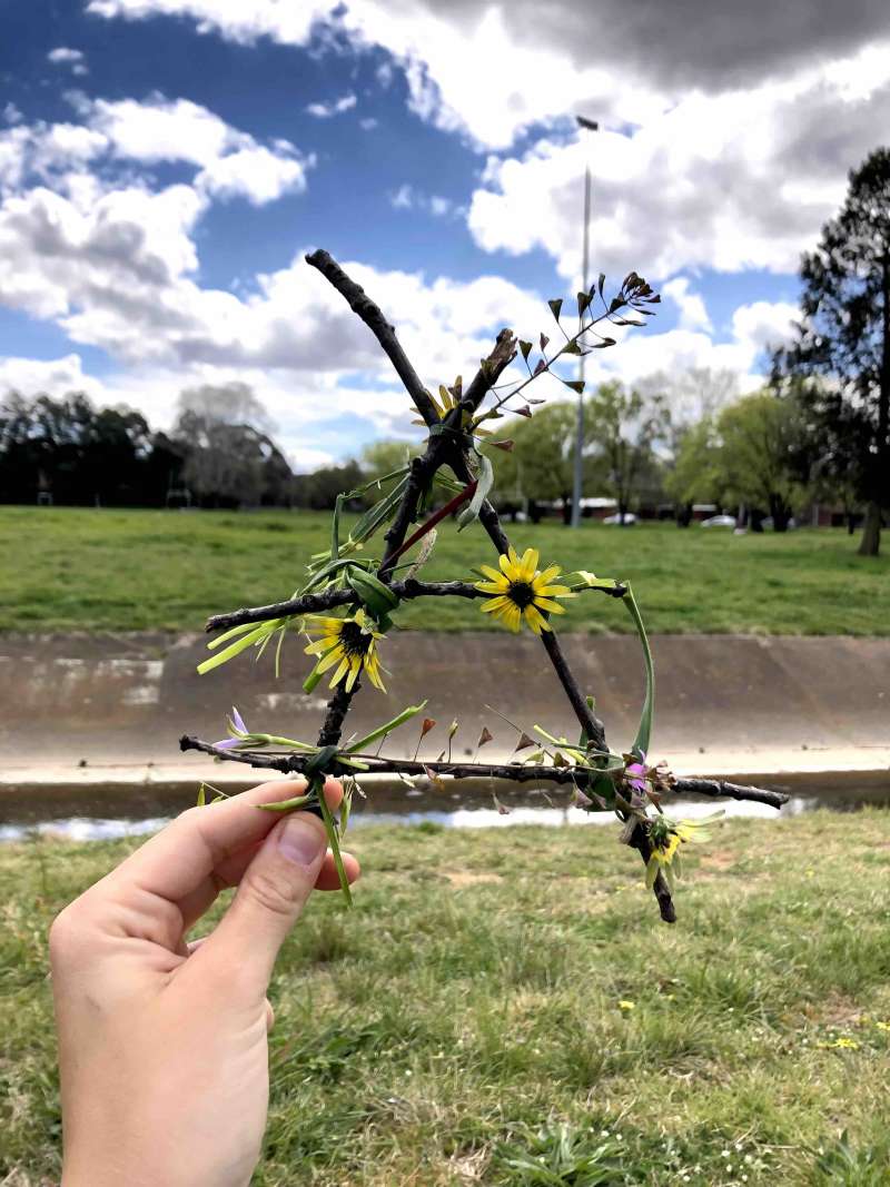 A close up of 4 sticks bound together to form a triangle shape with grass and flowers on it. A close up of 4 sticks bound together to form a triangle shape with grass and flowers on it.