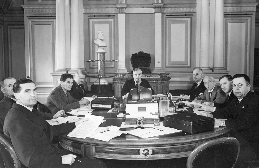 Nine men are gathered, seated around a large table covered with paper documents, with Robert Menzies seated at the head of the table. Nine men are gathered, seated around a large table covered with paper documents, with Robert Menzies seated at the head of the table.