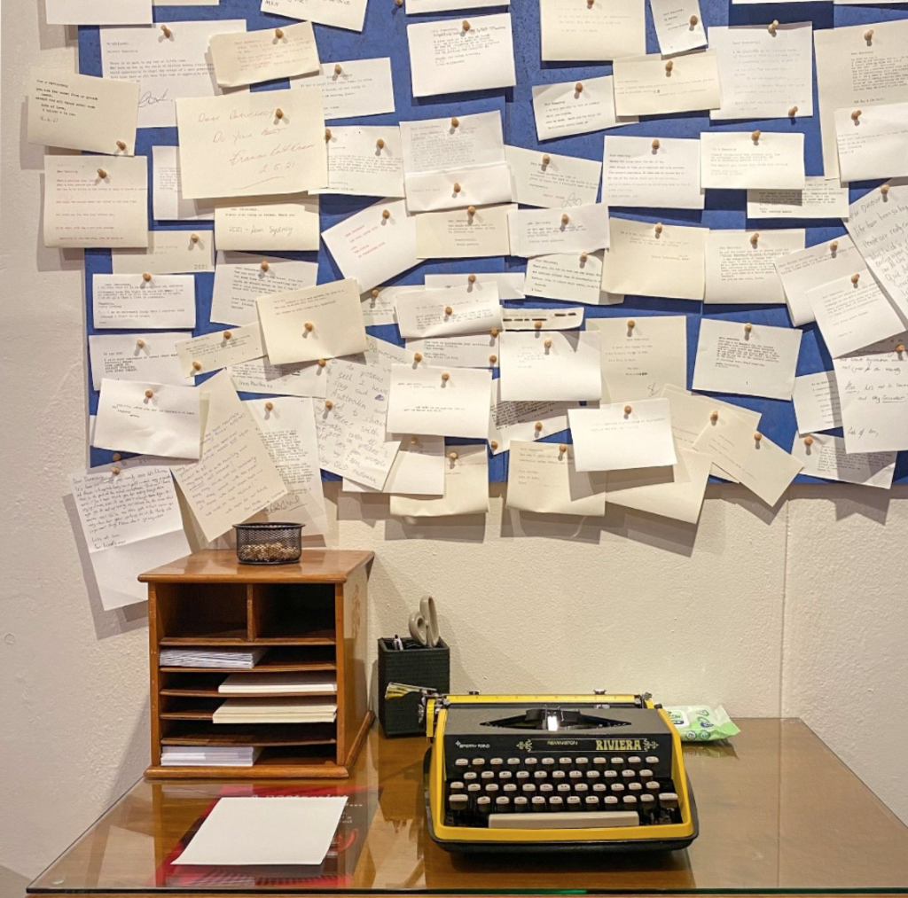 A yellow typewriter sits on a desk in front of a pinboard covered in typewritten notes. A yellow typewriter sits on a desk in front of a pinboard covered in typewritten notes.
