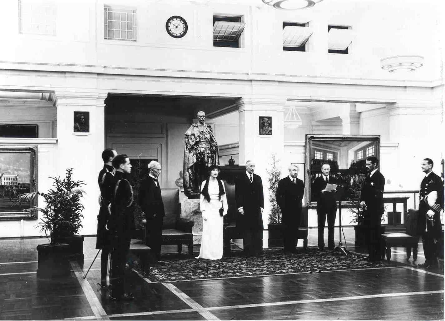 Governor-General Gowrie announces the abdication of King Edward VII and the accession of King George VI. This announcement occurred at 1.50am local time due to the time difference between London and Canberra.
National Library of Australia This black and white photograph shows a group of people standing at attention in King’s Hall while the announcement is read. The statue of George the Fifth looms over the group. Ther are seats behind the attendees, including the Vice Regal Chair which would have been moved from the Senate Chamber. The men wear dark suits or military uniforms while the Governor-General’s wife, Lady Gowrie, wears a white formal dress, hat and gloves.