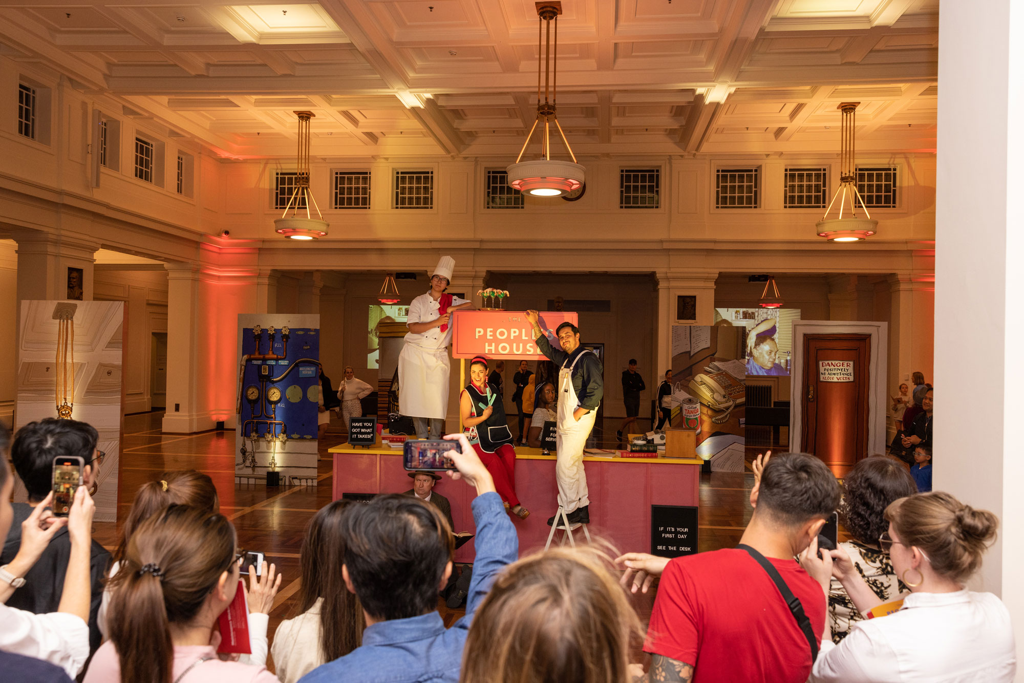 Visitors gather in King's Hall for the Enlighten festival in 2023. A group of visitors hold up their phones and film a performance with actors dressed as a chef, a hairdresser and a handyman inside King's Hall with a sign in the background that says The People's House.