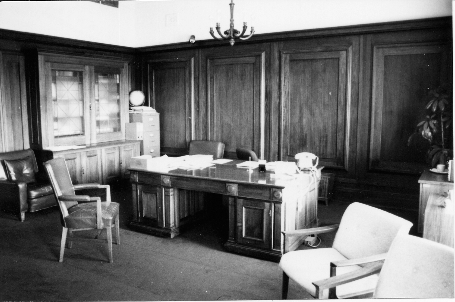 The Clerk of the Senate's office in 1985. It featured a substantial desk, built-in bookshelves and numerous mismatched chairs.
Photograph by Lori Anglin, Tanner and Associates, MoAD Collection This black and white photograph shows the layout of the Clerk of the Senate’s office in 1985 when it was occupied by Alan Cumming Thom. The timber-panelled room contains a large desk (originally designed by the building’s architect, John Smith Murdoch), glass-fronted built-in bookshelves and a sideboard with an indoor plant. There are a range of visitor chairs and an easy chair in the foreground.