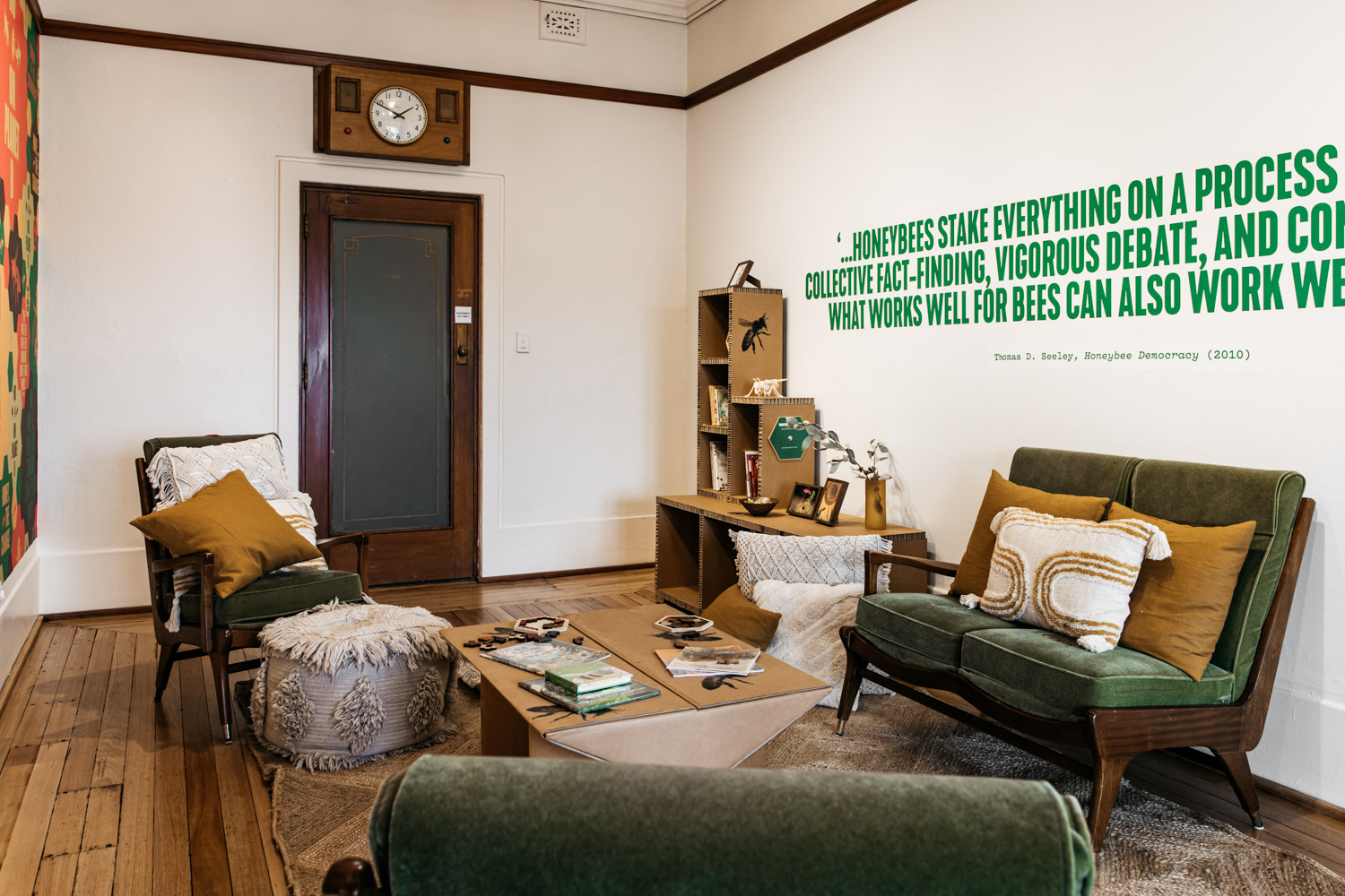 A room with green couches, cushions on the floor and books on the bookshelf.