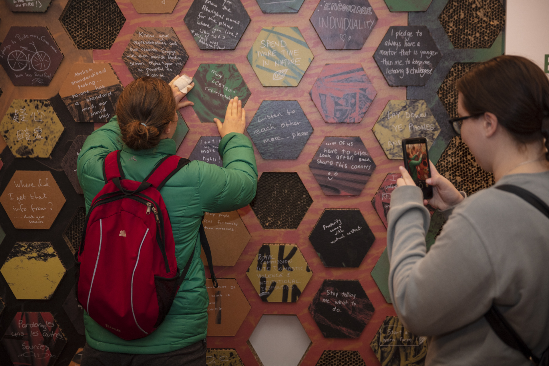 A woman takes a photo with her mobile photo of her friend touching a wall with colourful hexagons fitting into a wall.