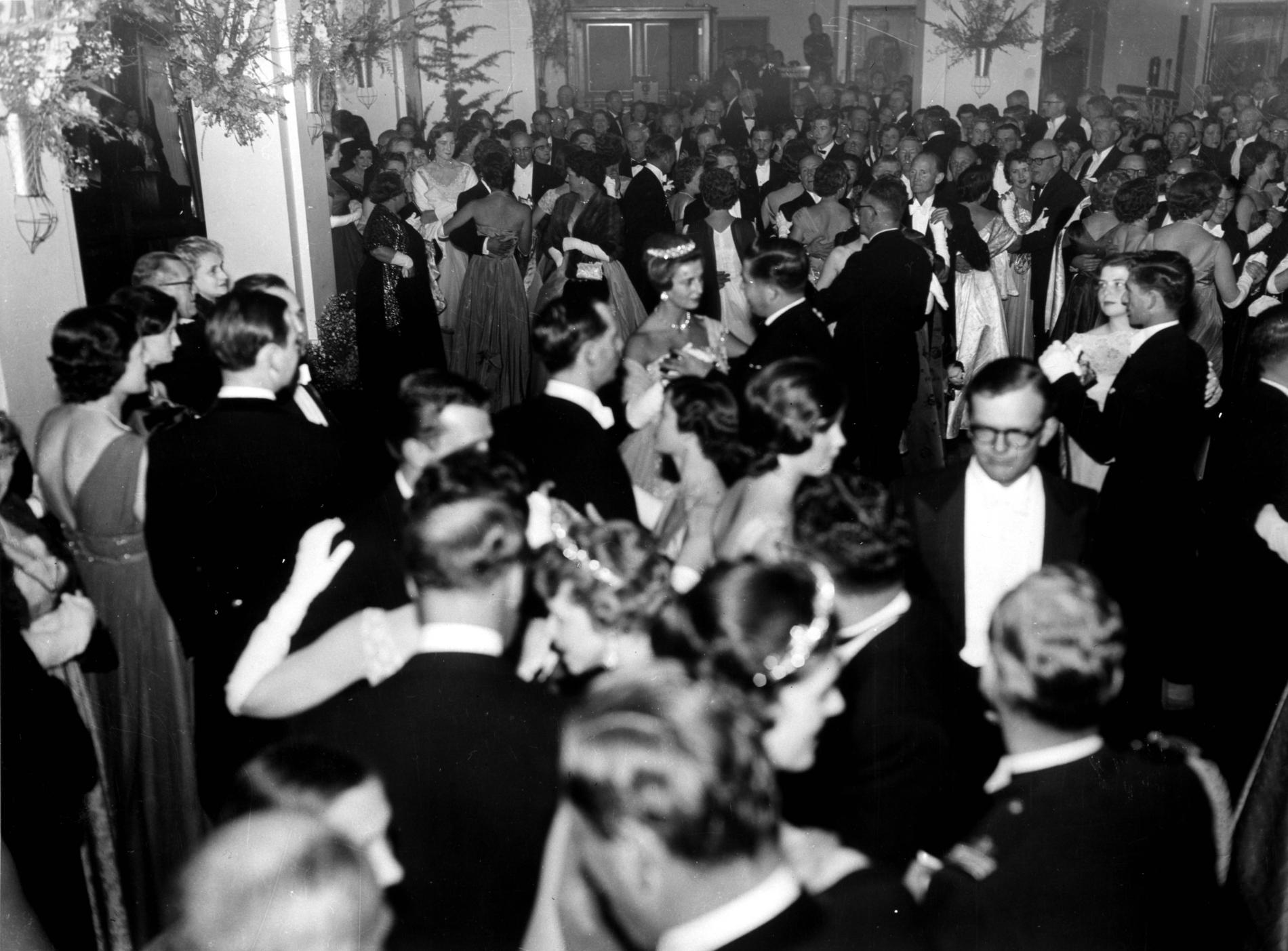 A ball is held in King's Hall for the visit of Princess Alexandra of Kent in 1959. Tickets were offered to under 25s through a ballot to reduce the average age of the guest list.
Photograph courtesy of Barbara Smith, Museum of Australian Democracy Collection In this black and white photograph a beautifully decorated King’s Hall is packed with people. In the background and left of the frame people stand and chat on the edge of the dance floor while those in the centre of the image and on the right are dancing what appears to be a waltz. Princess Alexandra in a sparkling tiara is prominent in the centre of the crowd. The men are in tuxedos and the women wear evening dresses with elbow length white gloves.