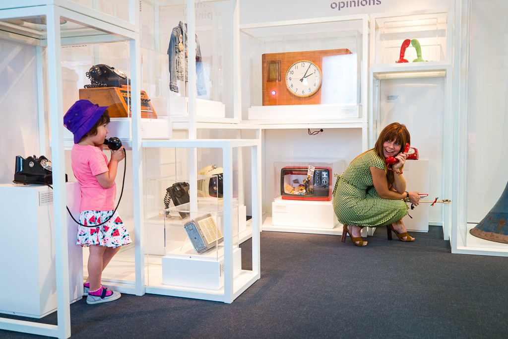 A child speaks on a phone talking to their mother surrounded by object cases in a museum with a clock, a radio and a TV.