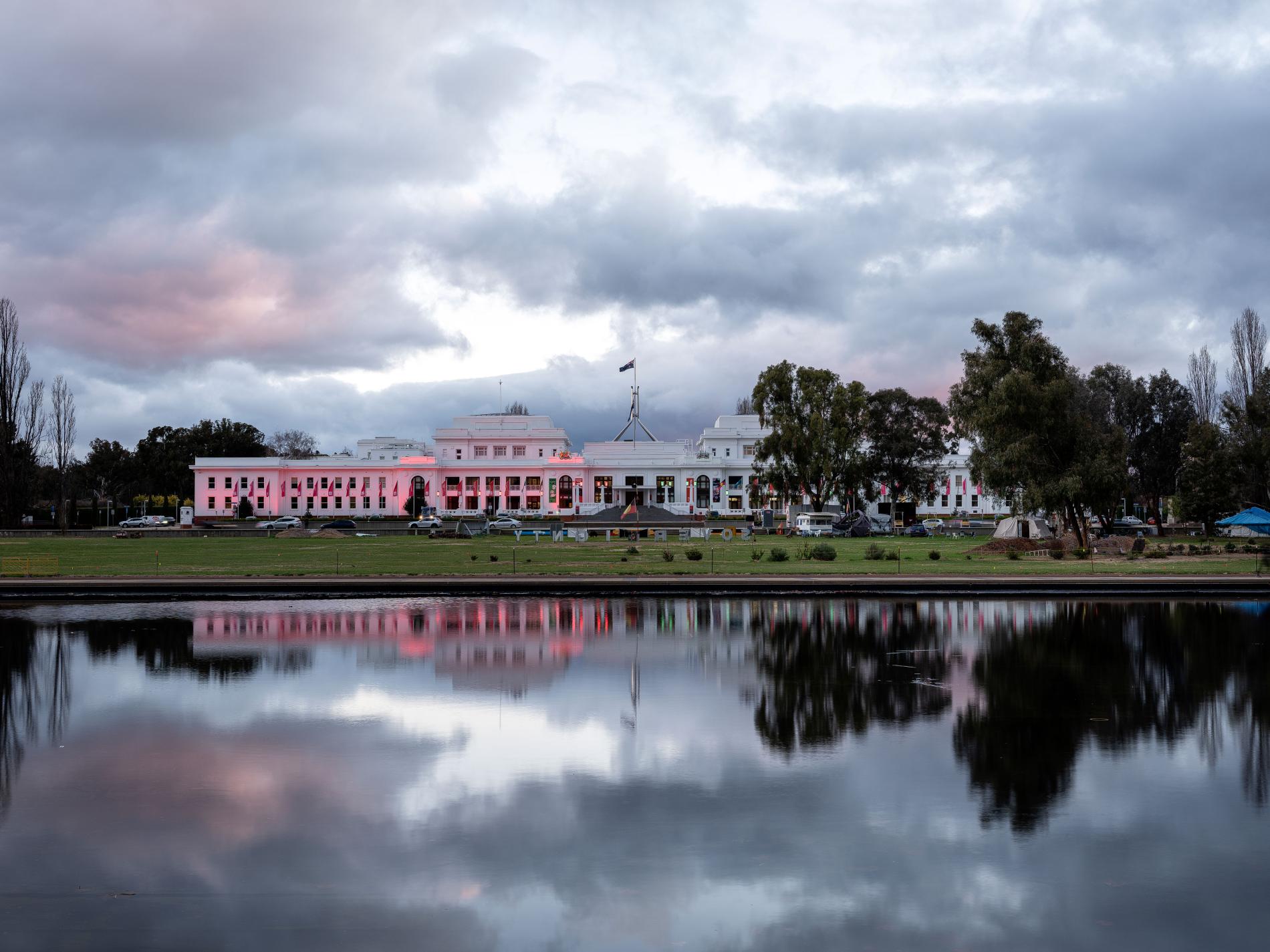 A large white building with the Australian flag on top, with a body of water in front that shows the reflection. The image is at dusk. A large white building with the Australian flag on top, with a body of water in front that shows the reflection. The image is at dusk.