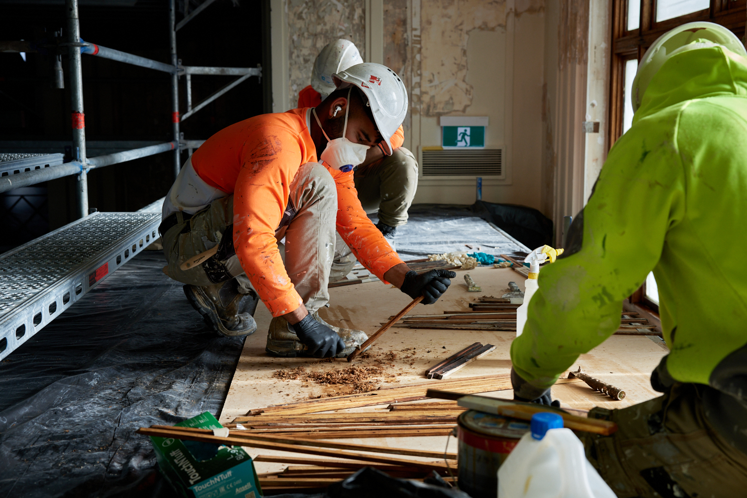 A construction working wearing a mask, an orange top and a hard hat kneels down to chip away at a wood panel below his feet.