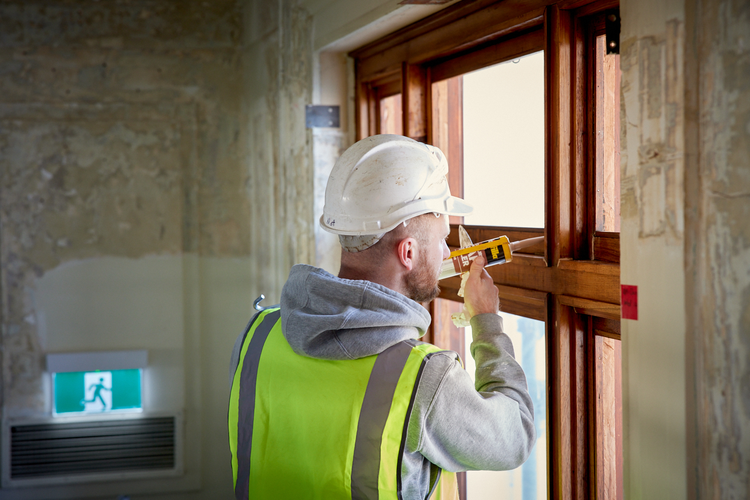 A construction working wearing a yellow hi-vis jacket and a help holds a glue gun up to a timber window frame.