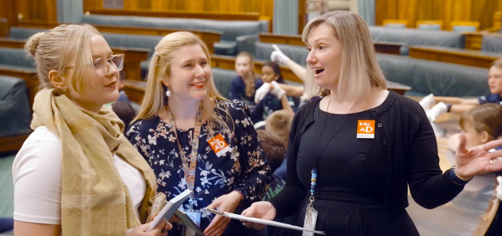 Two people stand holding clipboards, smiling and talking to a person about old parliament house's chamber of representatives. Two people stand holding clipboards, smiling and talking to a person about old parliament house's chamber of representatives.