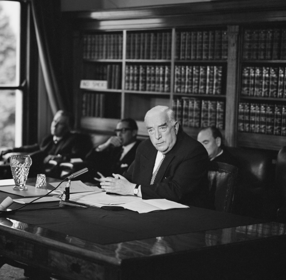 This black and white photograph shows Prime Minister Robert Menzies seated at a large table in the Government Party Room in 1965. He is conducting a press conference and there is a microphone, jug and water glass, and papers on the table. Behind him are three men seated in easy chairs and a bookcase filled with Hansards (volumes of parliamentary debates).  
