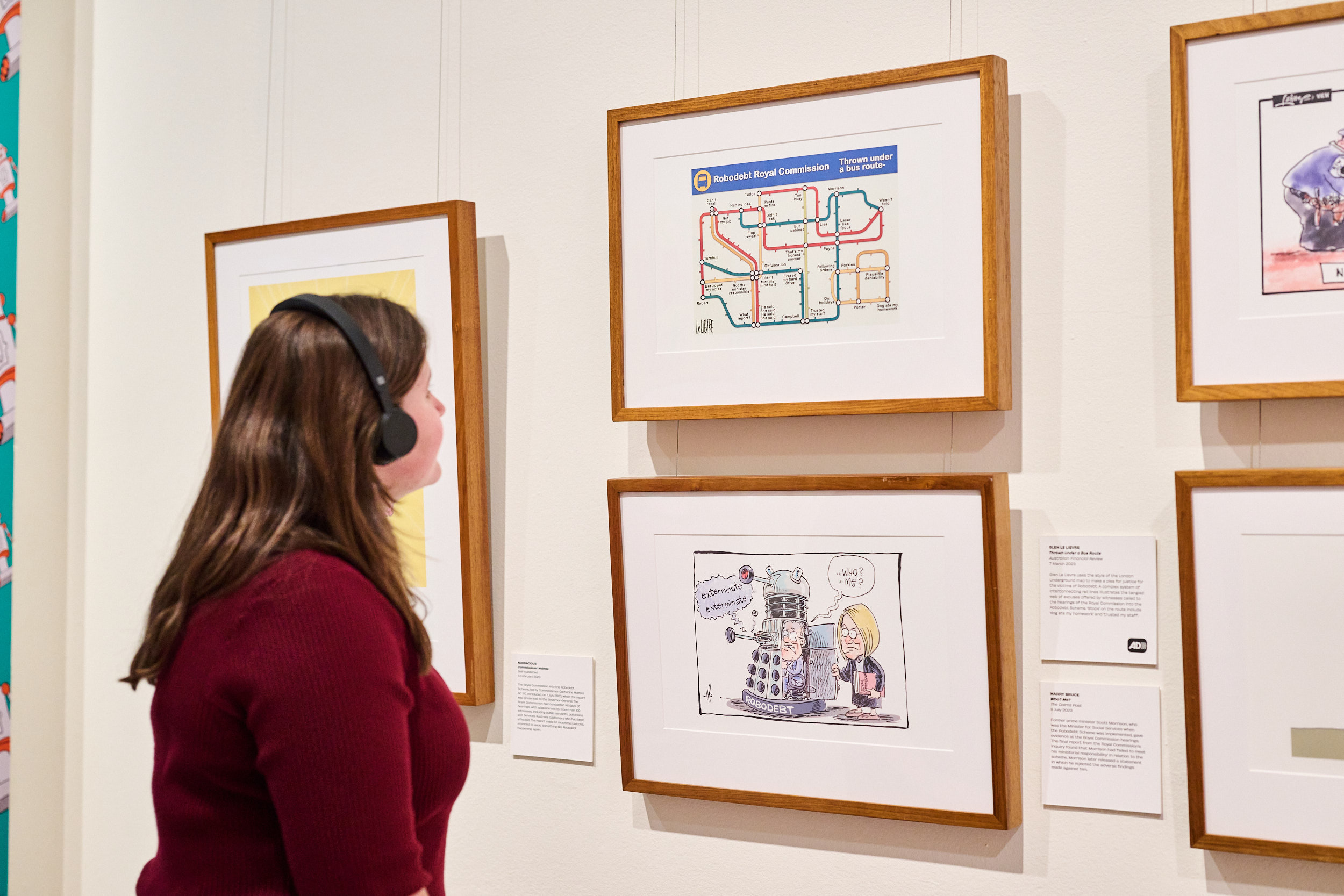 A woman wearing a dark red top and black headphones looks at framed cartoons with a caption featuring the Audio Description logo. A woman wearing a dark red top and black headphones looks at framed cartoons with a caption featuring the Audio Description logo.