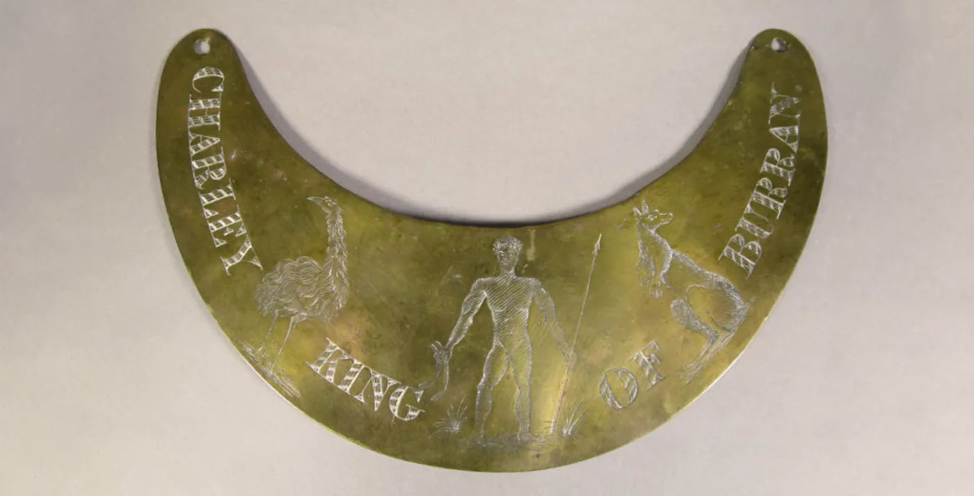 A brass breastplate with the words 'CHARLEY KING OF BURRAN' etched with illustrations of an emu, kangaroo, and a man holding a spear and two burren (boomerangs). A brass breastplate with the words 'CHARLEY KING OF BURRAN' etched with illustrations of an emu, kangaroo, and a man holding a spear and two burren (boomerangs).