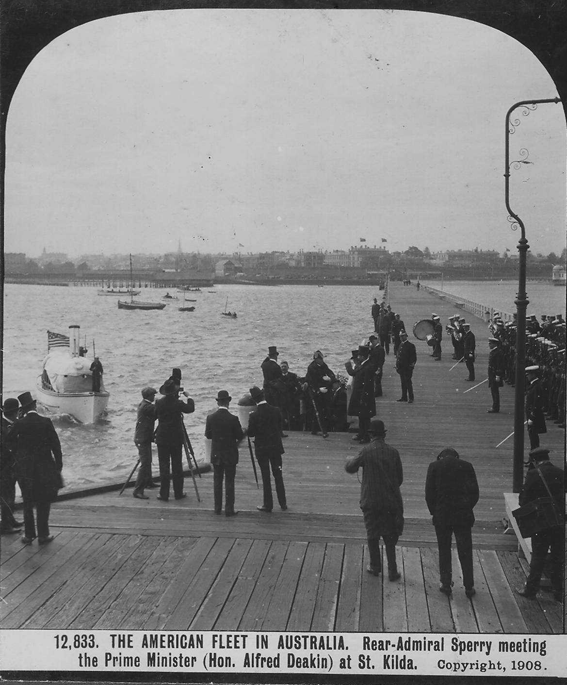 An elevated view from above and behind small clusters of people who stand on a wooden pier. They are looking out at a boat that is headed towards the pier. The label reads "The American Fleet in Australia. Rear-Admiral Sperry meeting the Prime Minister (Hon. Alfred Deakin) at St. Kilda." 1908. An elevated view from above and behind small clusters of people who stand on a wooden pier. They are looking out at a boat that is headed towards the pier. The label reads "The American Fleet in Australia. Rear-Admiral Sperry meeting the Prime Minister (Hon. Alfred Deakin) at St. Kilda." 1908.