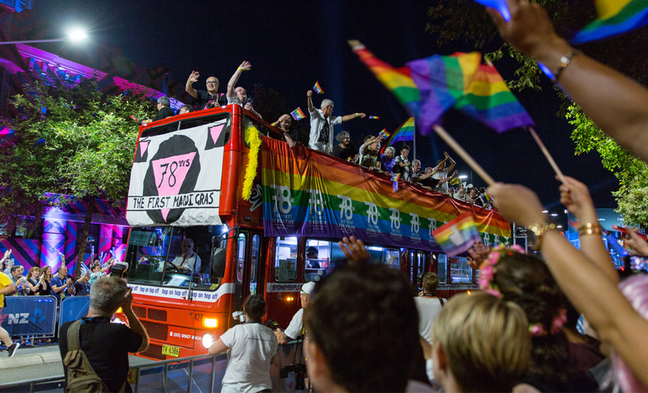 A red double-decker bus with rainbow banners displaying the words '78ers: the first mardi gras' with people on top of the bus waving to crowds on each side of the street. A red double-decker bus with rainbow banners displaying the words '78ers: the first mardi gras' with people on top of the bus waving to crowds on each side of the street.