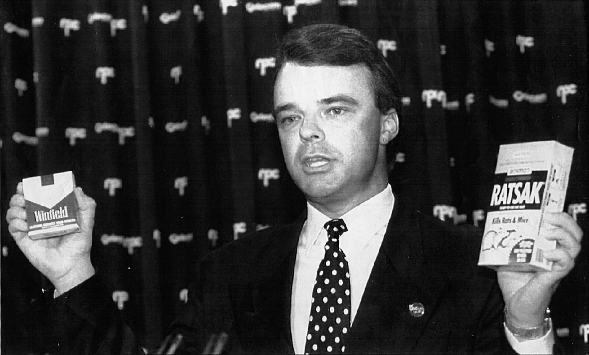 Dr Brendan Nelson, wearing a suit and spotted tie, holding a 'Winfield' cigarette box in one hand and a 'Ratsak' box in the other. Dr Brendan Nelson, wearing a suit and spotted tie, holding a 'Winfield' cigarette box in one hand and a 'Ratsak' box in the other.