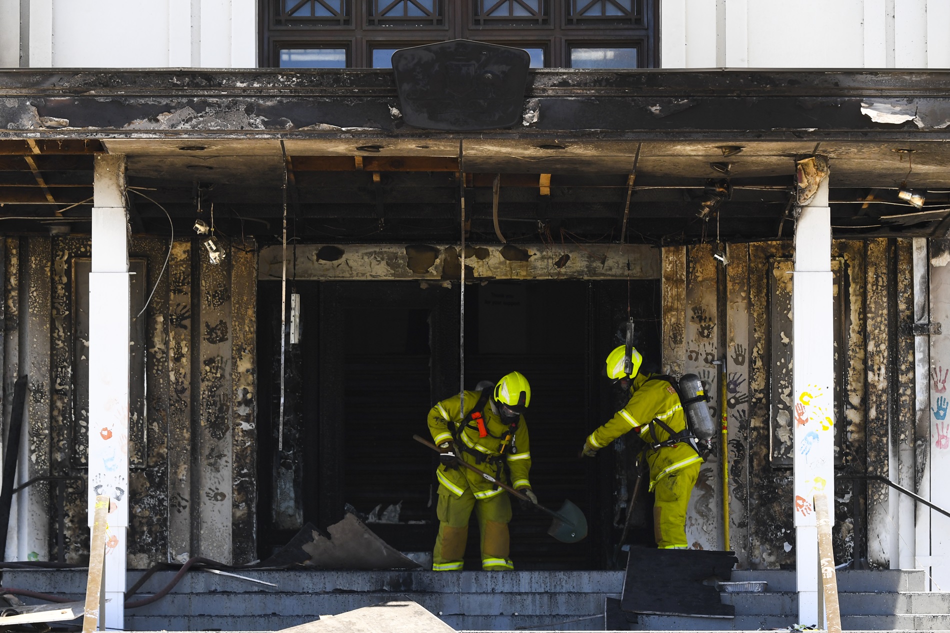 Two firefighters dressed in yellow hi-vis clothing and protective equipment, hold shovels and assess the fire damage to the front entrance of Old Parliament House. Two firefighters dressed in yellow hi-vis clothing and protective equipment, hold shovels and assess the fire damage to the front entrance of Old Parliament House.