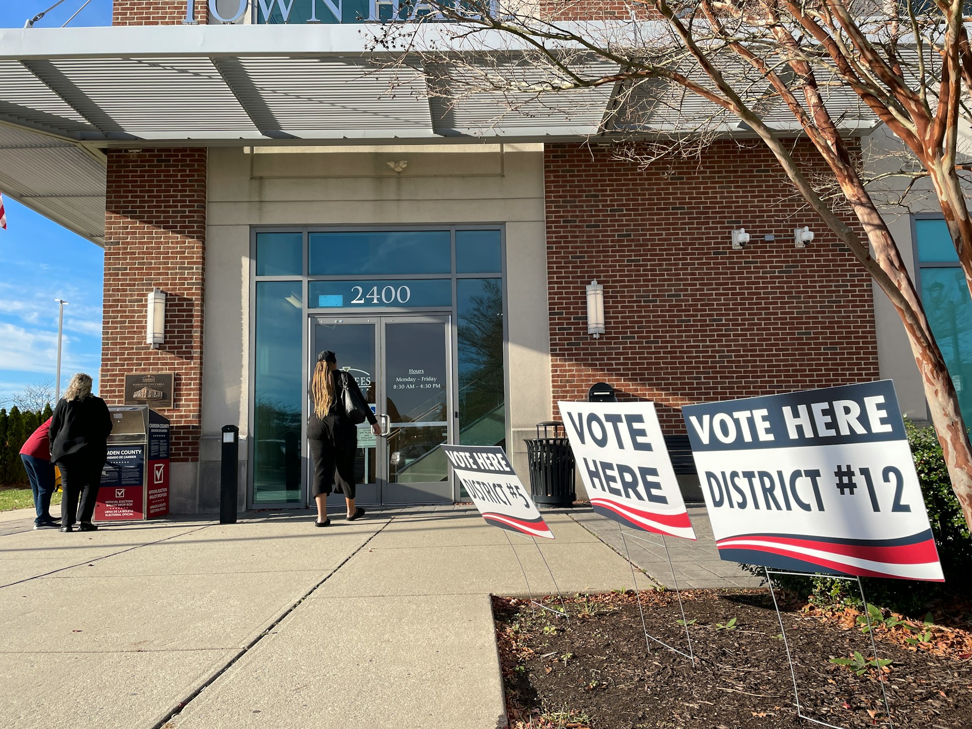 The front of a building with signage that reads 'Vote here', 'District #5' and 'District #12'. One person enters the building, whilst two others gather around a box that reads 'Official election ballot drop box'. The front of a building with signage that reads 'Vote here', 'District #5' and 'District #12'. One person enters the building, whilst two others gather around a box that reads 'Official election ballot drop box'.