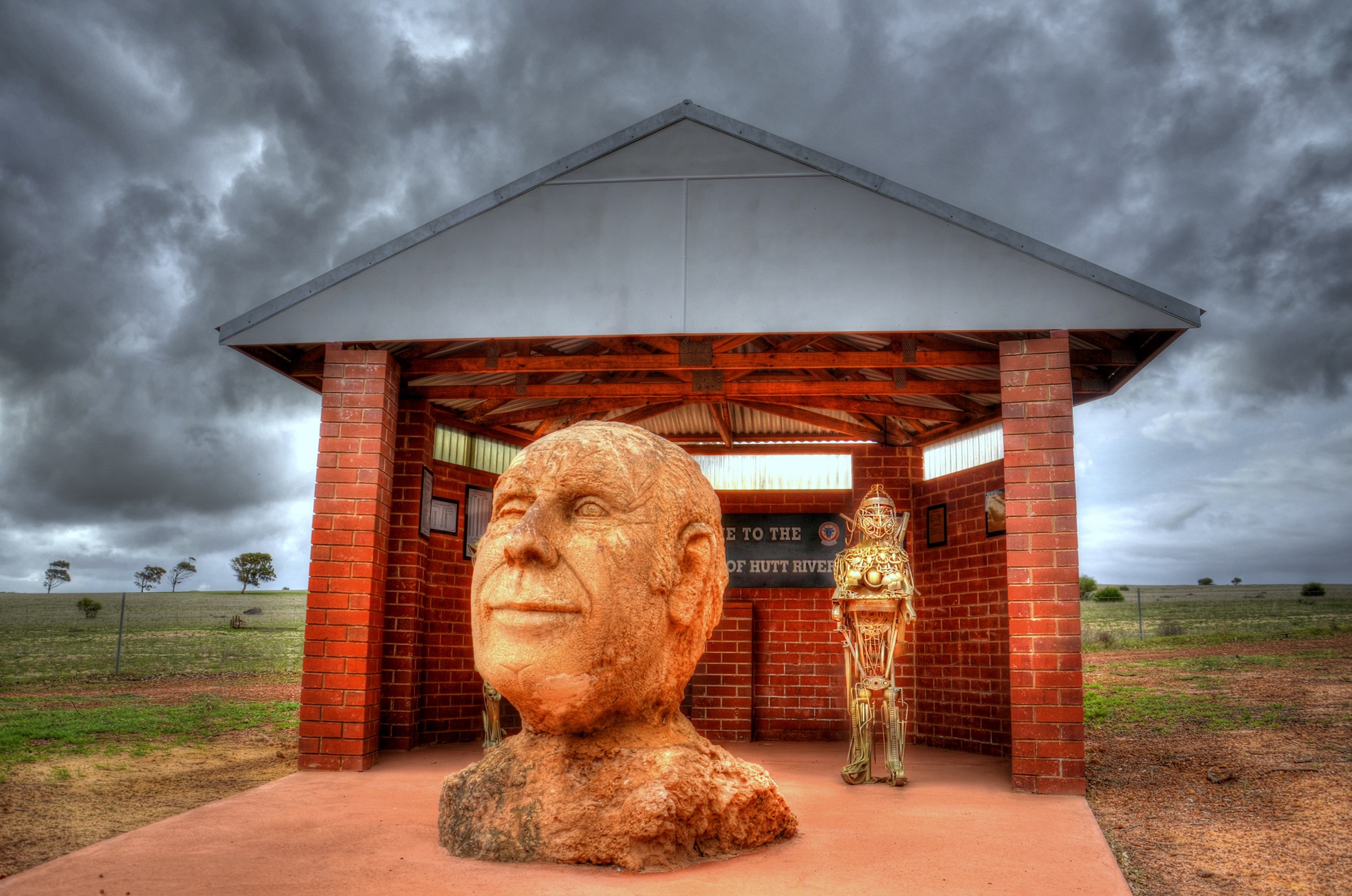 A hand-carved rock bust depicting the head and shoulders of 'Prince' Leonard George Casley, positioned in front of a redbrick shelter in a paddock. A hand-carved rock bust depicting the head and shoulders of 'Prince' Leonard George Casley, positioned in front of a redbrick shelter in a paddock.