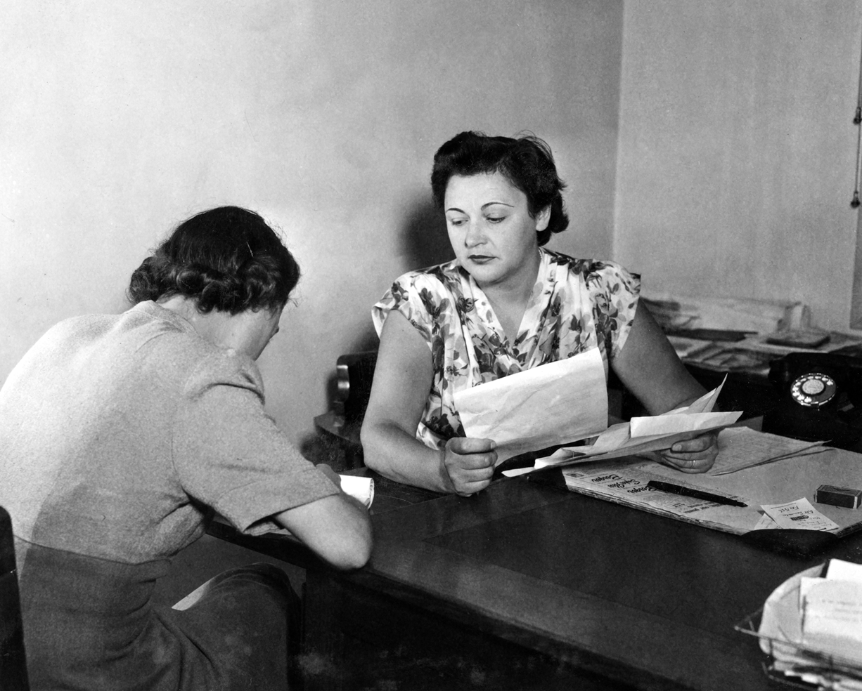Nancy Wake is at a desk holding papers. She is looking across at another woman who is writing. Nancy Wake is at a desk holding papers. She is looking across at another woman who is writing.