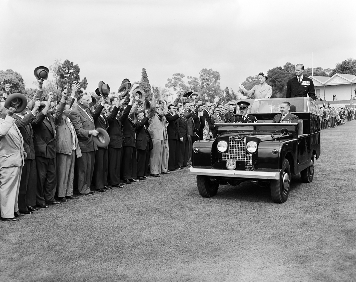 The Queen and Prince Phillip are standing in the back of a car waving to a crowd who are all waving their hats to them. The Queen and Prince Phillip are standing in the back of a car waving to a crowd who are all waving their hats to them.