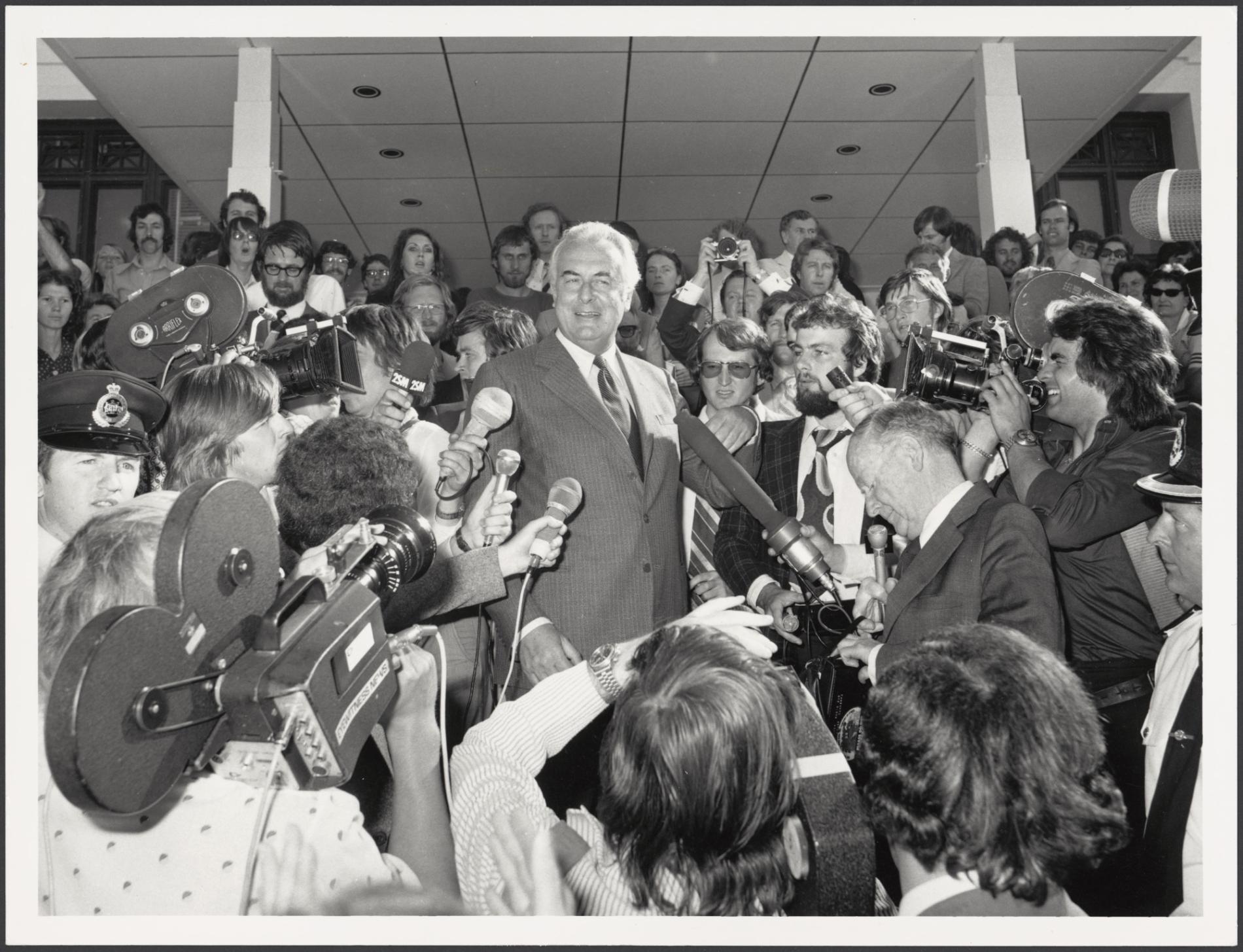 Gough Whitlam on the steps of Old Parliament House surrounded by the press who are holding microphones and video cameras on 11 November 1975.