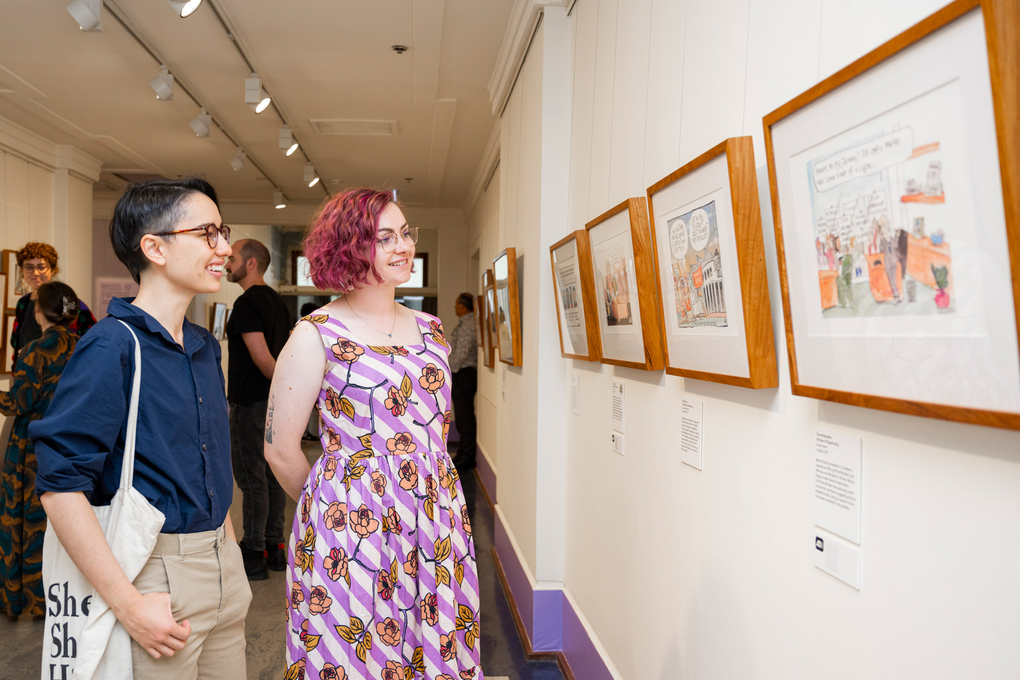 Two smiling people look at framed cartoons on the wall of an exhibition. 