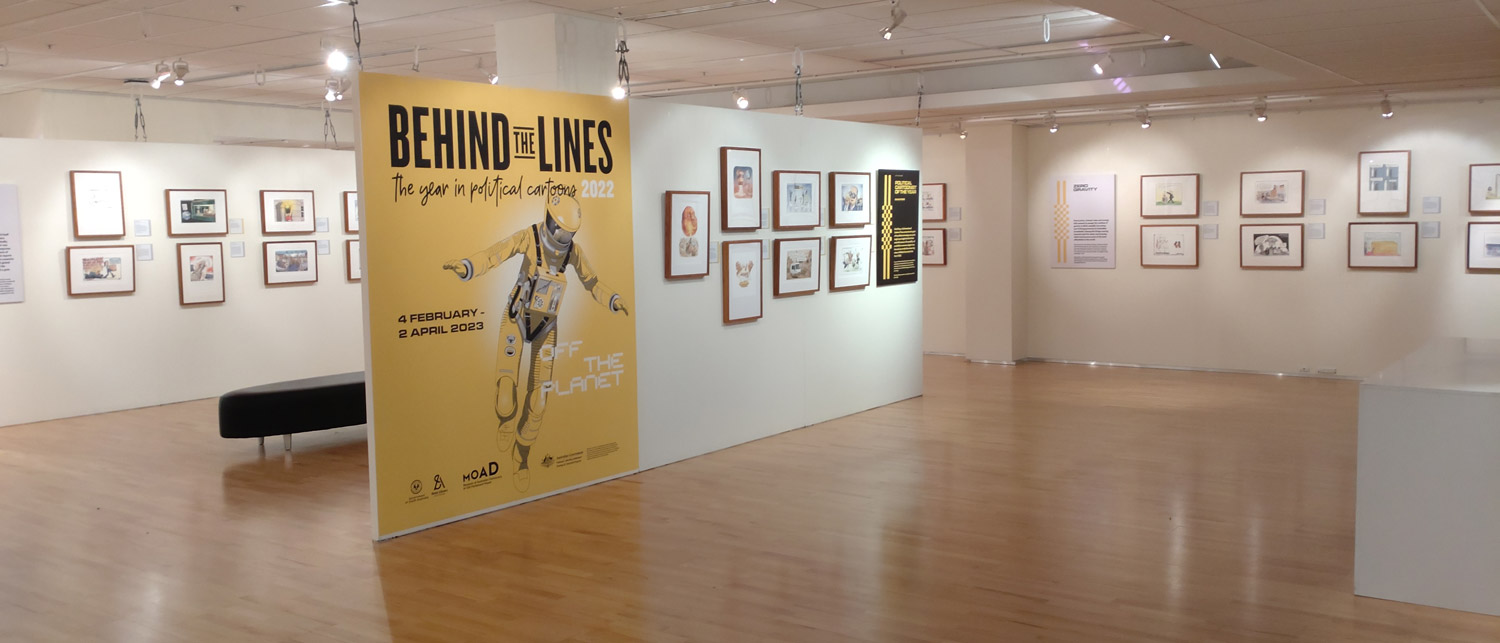 A gallery space with framed cartoons on the walls. A large, yellow introductory panel features the illustration of an astronaut and the words 'Behind the Lines'. A gallery space with framed cartoons on the walls. A large, yellow introductory panel features the illustration of an astronaut and the words 'Behind the Lines'.
