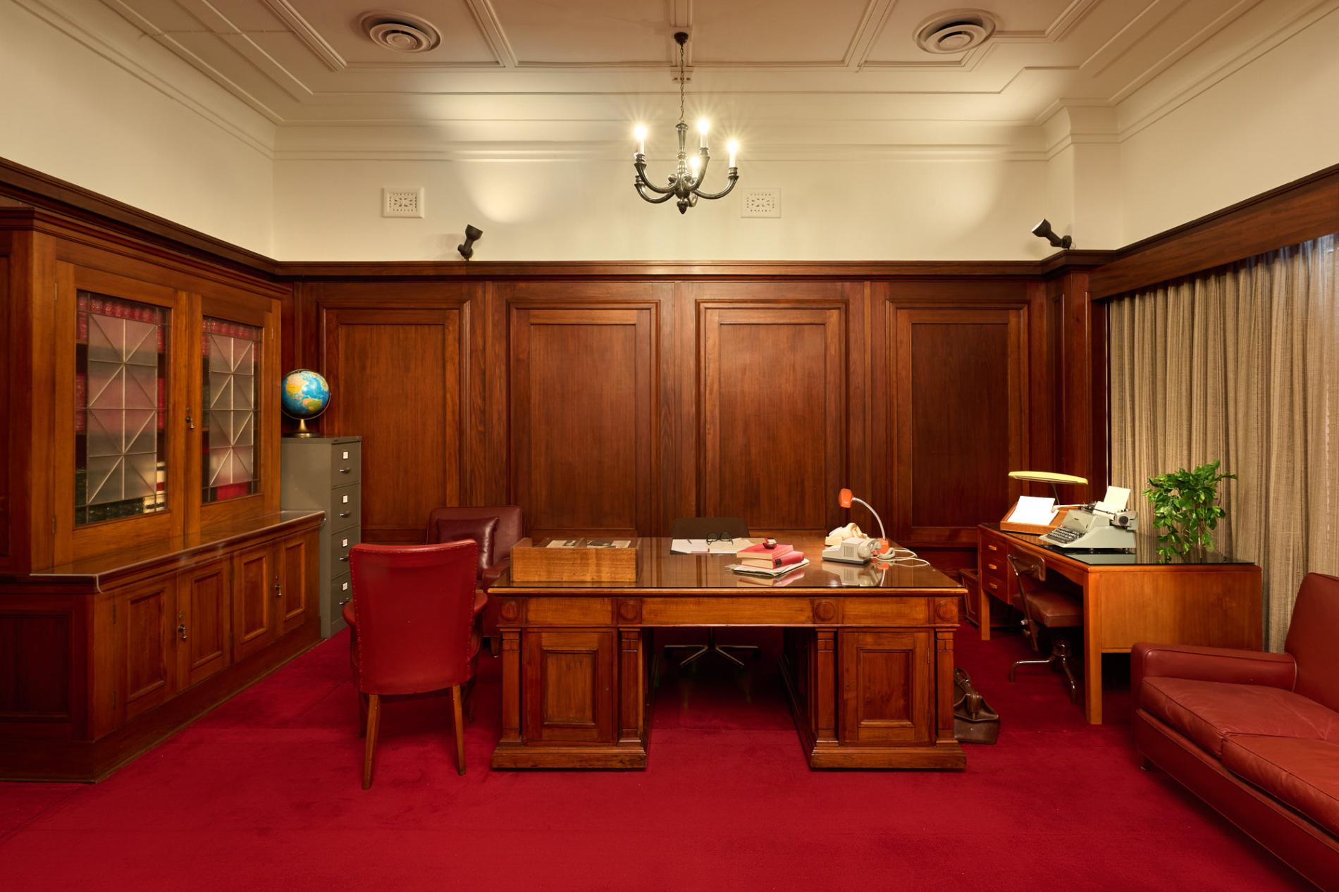 A wood-panelled room in Old Parliament House with two wooden desks, a wooden bookcase, red leather chairs and a red leather couch. The carpet is red.