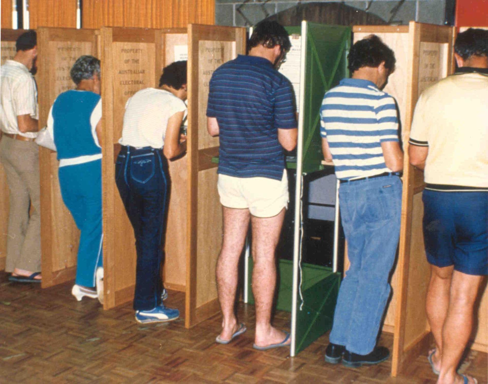 Men and women dressed in 1970s fashion vote in partitioned booths which are labelled 'Property of the Australian Electoral Office'.