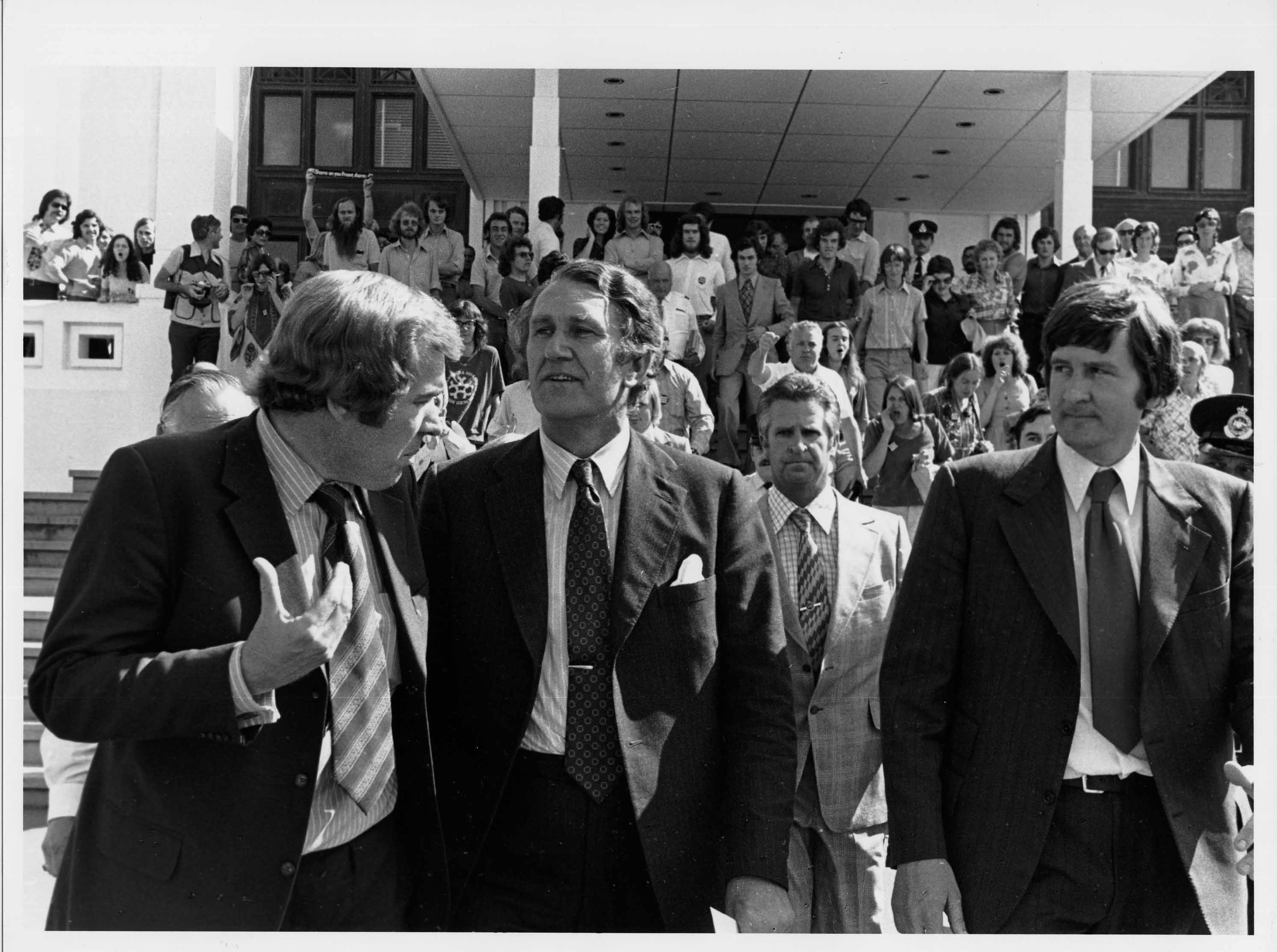 Malcolm Fraser walks away from Parliament House with two other men in suits. A crowd of people watch from the steps behind them. Malcolm Fraser walks away from Parliament House with two other men in suits. A crowd of people watch from the steps behind them.