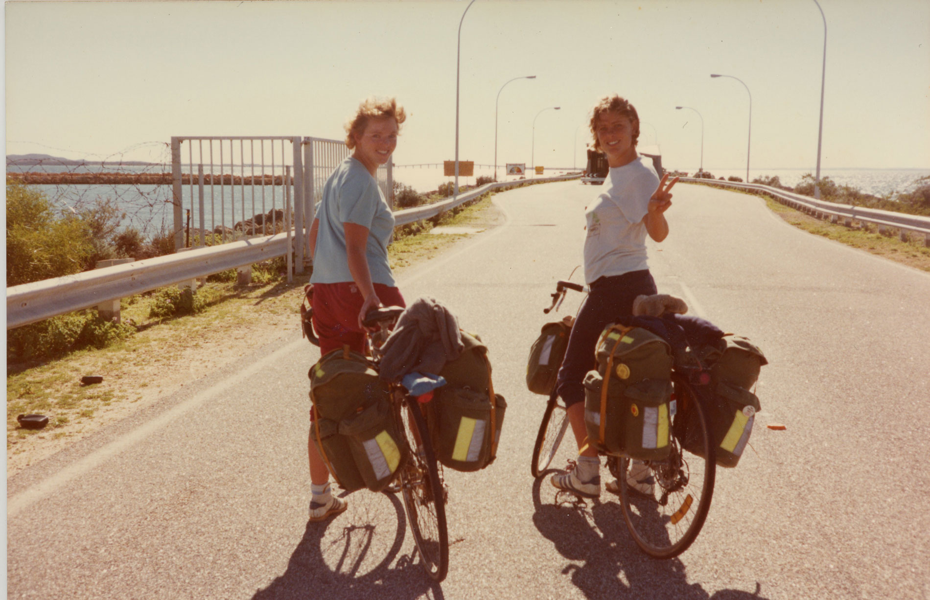 Two women on bikes on a road near the ocean.