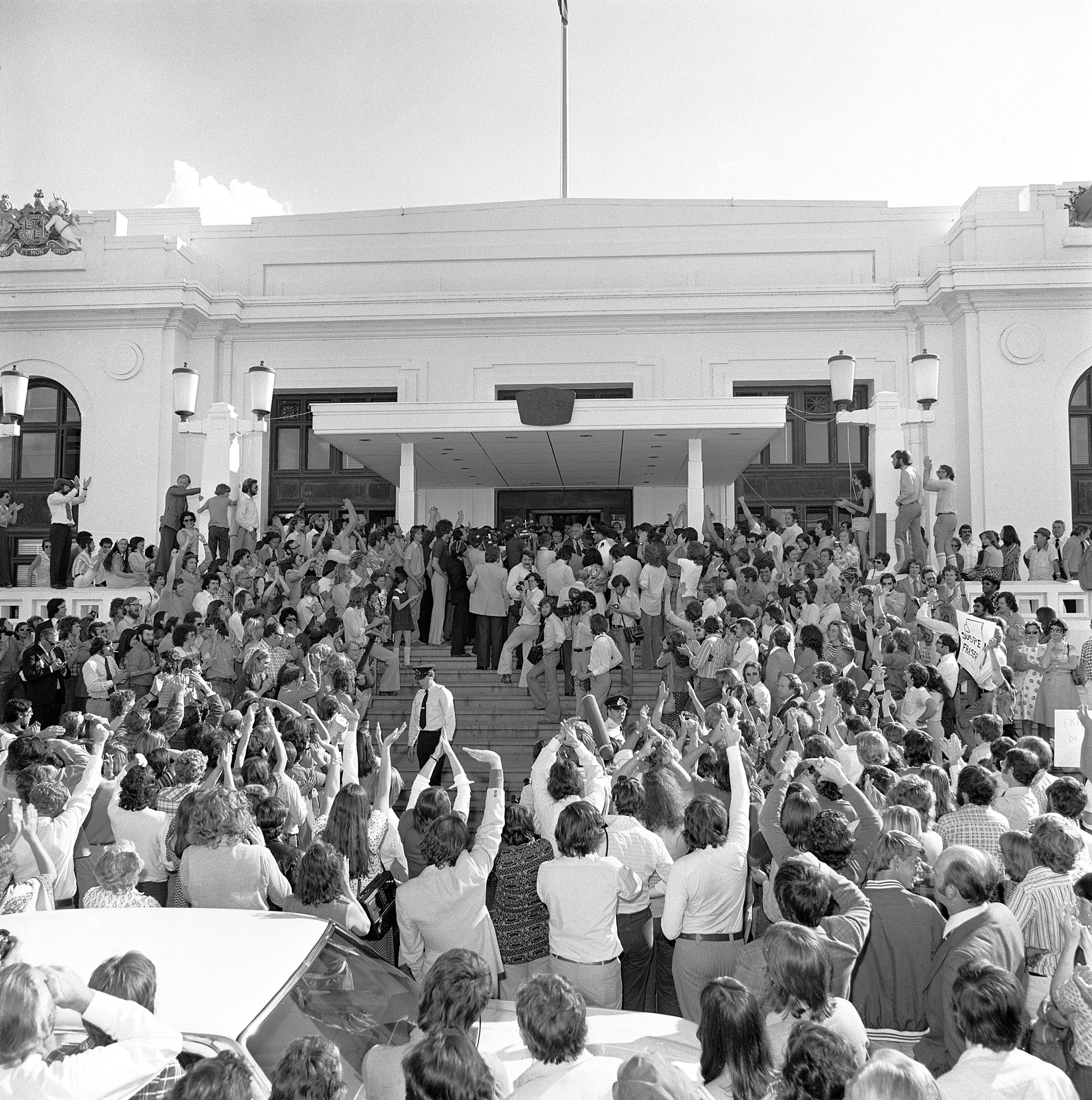 A large crowd of people and press stand below and on the steps of Parliament House after the dismissal of the Whitlam government.