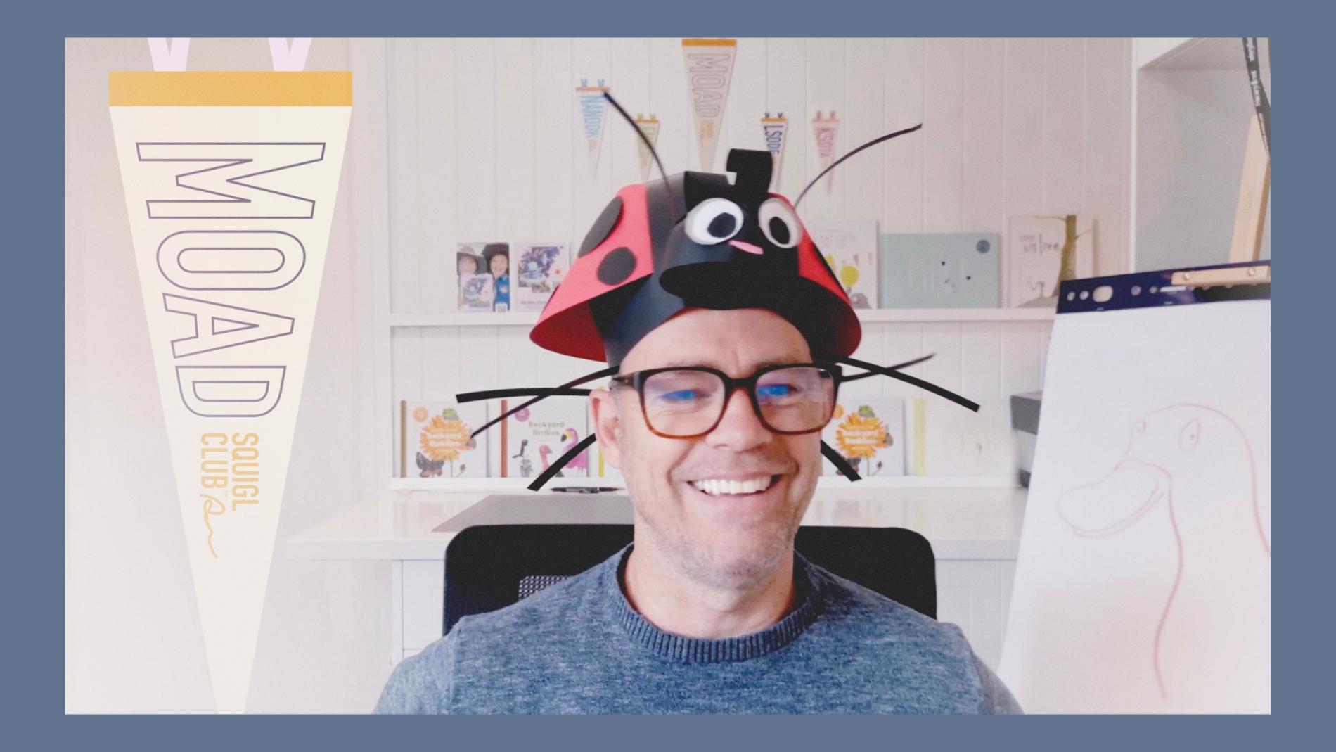 A man wearing a ladybug hat sits in front of a bookcase of children's books.