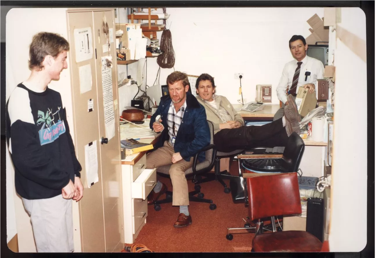 This amateur colour photograph gives us a peek into the small, crowded and messy office occupied by the Telecom employees at Old Parliament House. The office is crowded with desks, chairs, bookshelves and a cupboard. A young, smiling Scott Cadden stands to the left in jeans and sweatshirt, Neil Baker sits at a desk holding a telephone handset and dressed in trousers, checked shirt and casual blue jacket. He has red hair and a beard and was known as ‘Bluey’. Seated behind Neil is a smiling Jeff Wilke with hi This amateur colour photograph gives us a peek into the small, crowded and messy office occupied by the Telecom employees at Old Parliament House. The office is crowded with desks, chairs, bookshelves and a cupboard. A young, smiling Scott Cadden stands to the left in jeans and sweatshirt, Neil Baker sits at a desk holding a telephone handset and dressed in trousers, checked shirt and casual blue jacket. He has red hair and a beard and was known as ‘Bluey’. Seated behind Neil is a smiling Jeff Wilke with hi