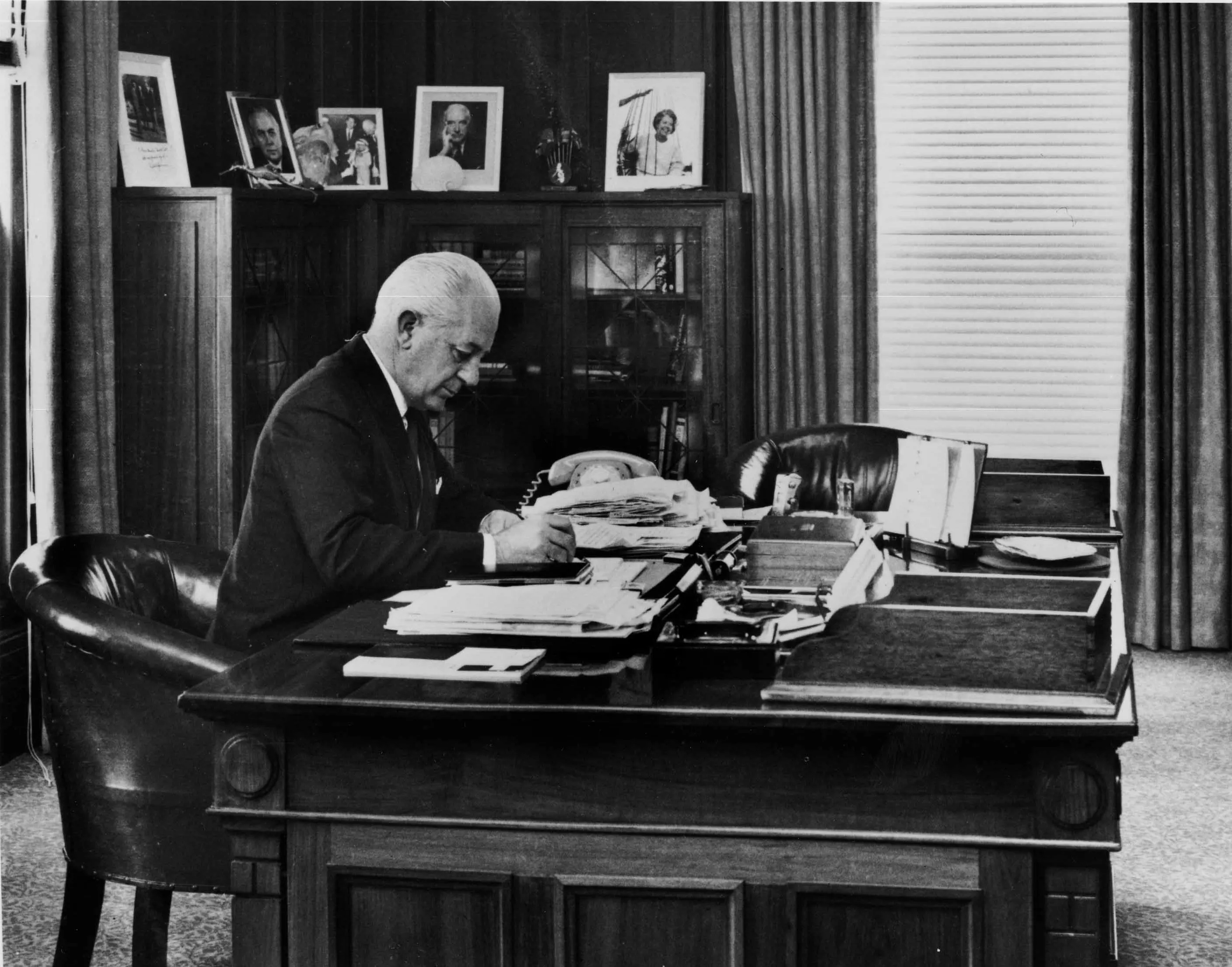 In this black and white photograph Prime Minister Harrold Holt sits at the timber desk made in 1926. Holt is wearing a dark suit, white shirt and tie and is leaning on the desk while he writes. The desk has fine carved panels with a geometric detailing on the corners. The surface of the desk is cluttered with stacks of documents and a telephone. On the glass-fronted cabinets in the background are a number of framed photographic portraits. In this black and white photograph Prime Minister Harrold Holt sits at the timber desk made in 1926. Holt is wearing a dark suit, white shirt and tie and is leaning on the desk while he writes. The desk has fine carved panels with a geometric detailing on the corners. The surface of the desk is cluttered with stacks of documents and a telephone. On the glass-fronted cabinets in the background are a number of framed photographic portraits.
