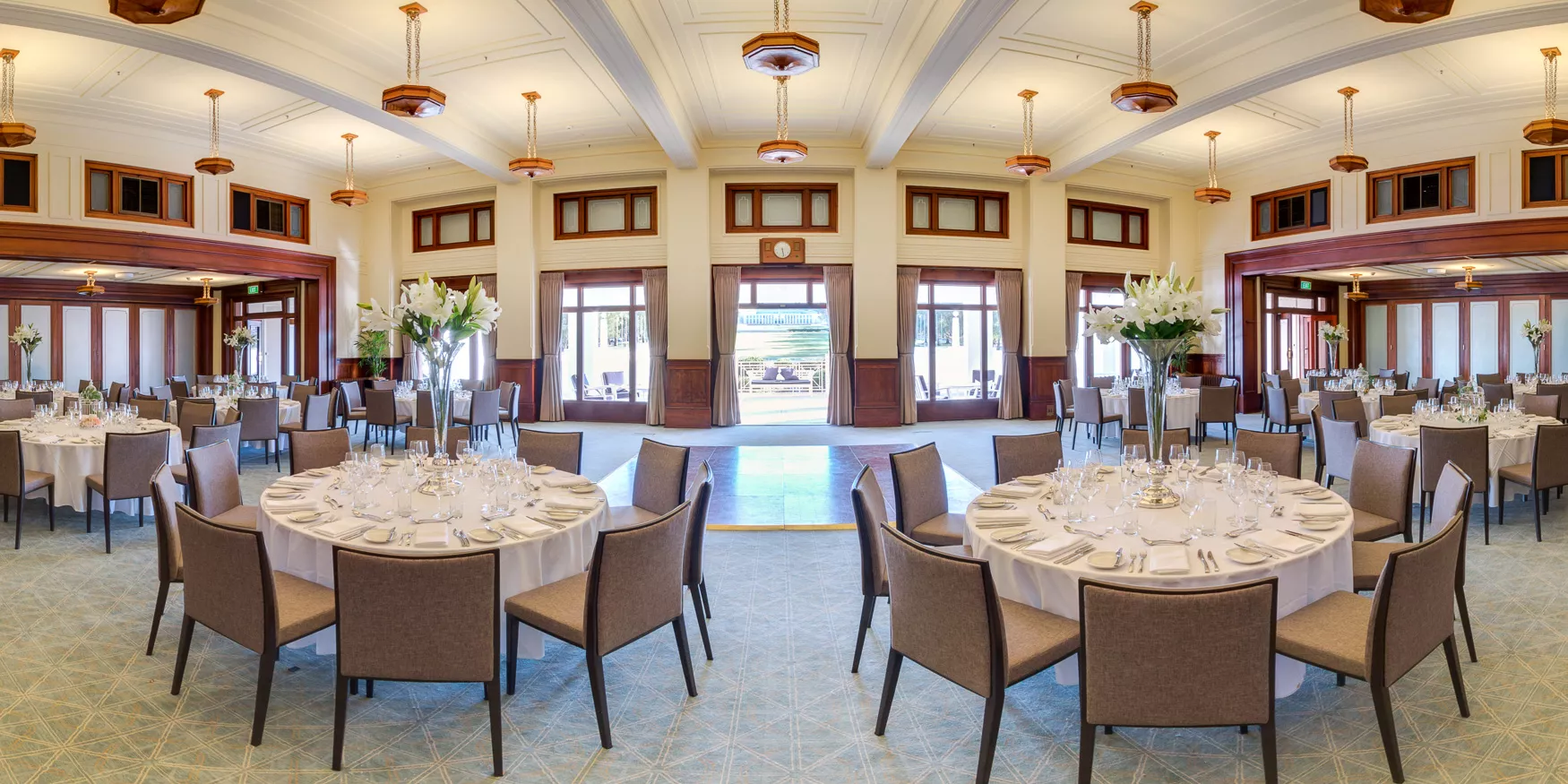 An art deco styled room with wooden pendant chandeliers, white ceilings and carpeted floor, with circular tables set with white tablecloths and lilies. An art deco styled room with wooden pendant chandeliers, white ceilings and carpeted floor, with circular tables set with white tablecloths and lilies.