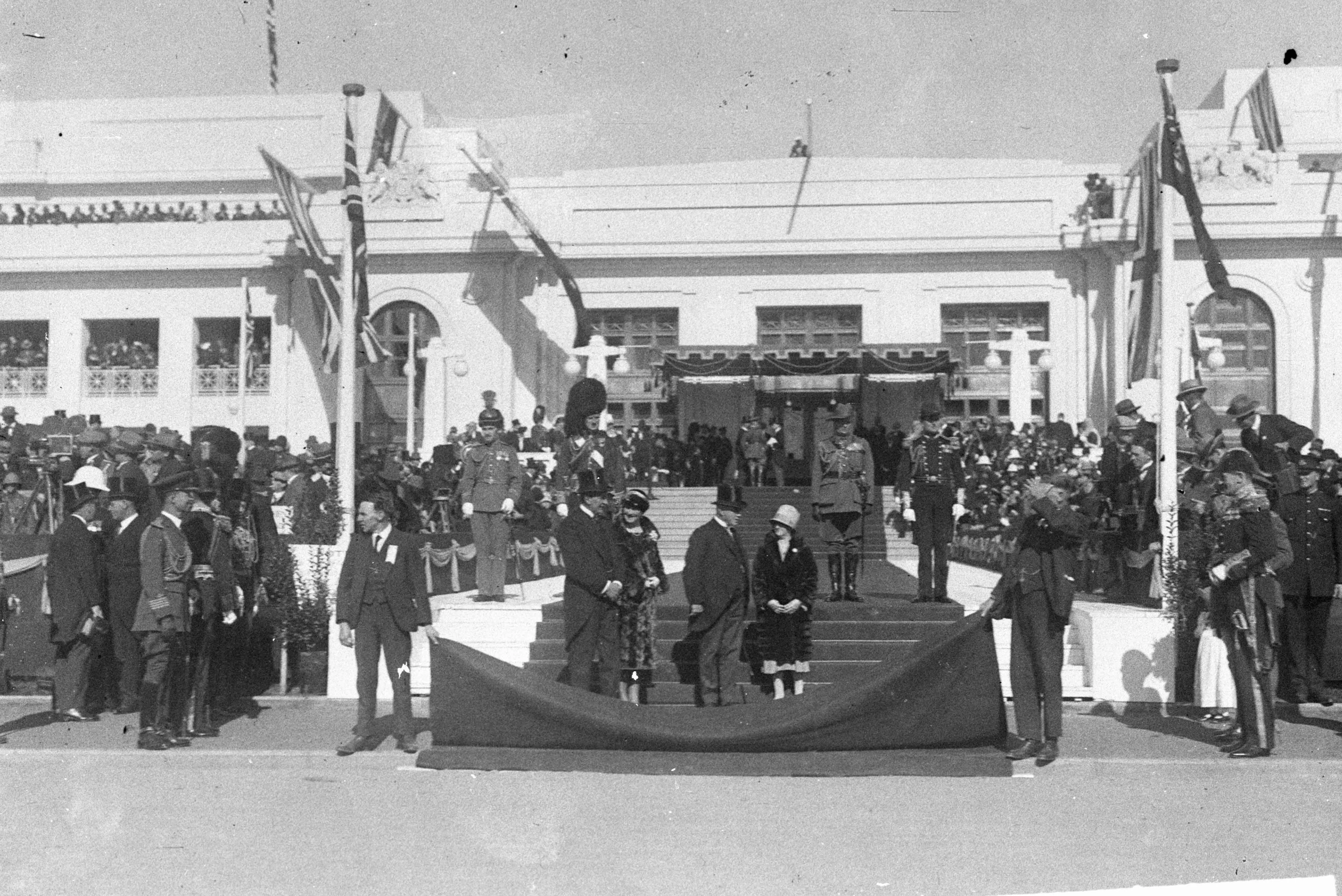 A black and white photo of the facade of Parliament House decorated with flags, a canopy and welcome carpet before the arrival of the Duke and Duchess of York for the opening of the building. There are crowds on the roof, front steps, verandahs and flanking the front door. A guard of honour in military uniform stands ready. Two attendants are holding up the end of the red carpet. A black and white photo of the facade of Parliament House decorated with flags, a canopy and welcome carpet before the arrival of the Duke and Duchess of York for the opening of the building. There are crowds on the roof, front steps, verandahs and flanking the front door. A guard of honour in military uniform stands ready. Two attendants are holding up the end of the red carpet.