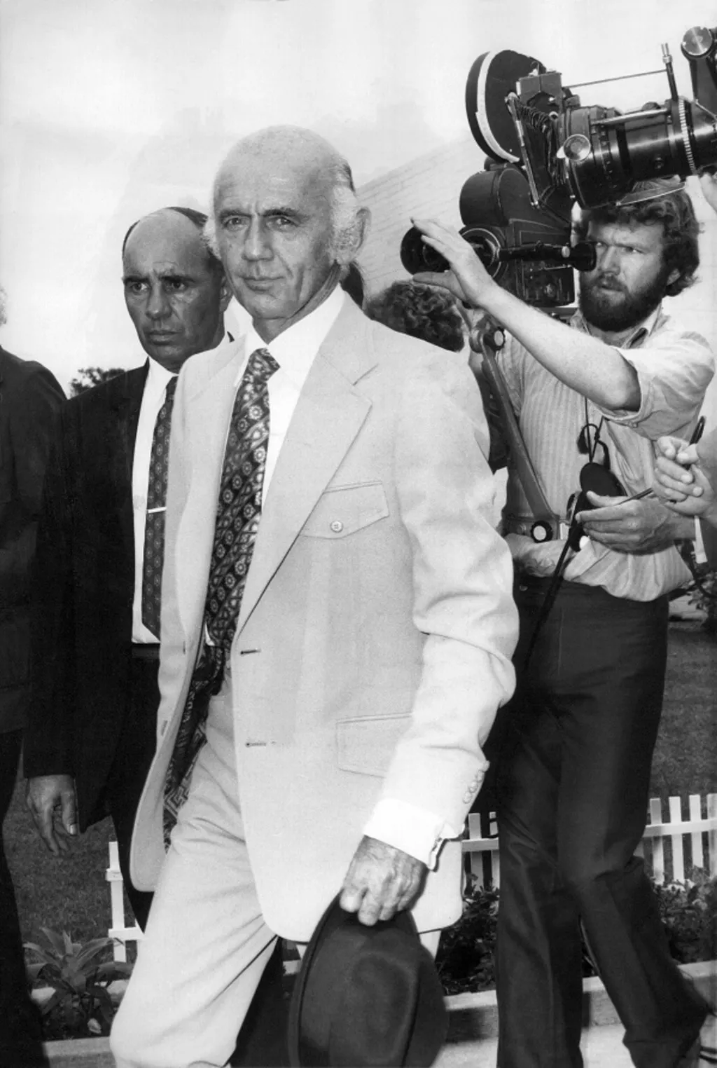 Billy McMahon wearing a light-coloured suit, hat in hand, followed by a film camera operator. Billy McMahon wearing a light-coloured suit, hat in hand, followed by a film camera operator.