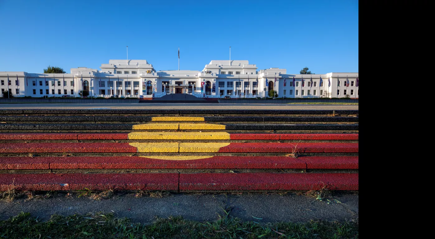 Steps with the Aboriginal flag painted on them lead up to a view of a large long white building, Old Parliament House.
