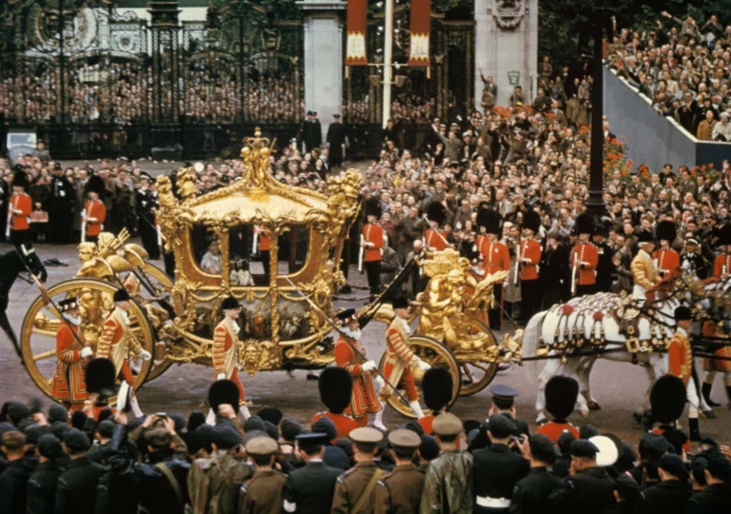 Queen Elizabeth II sits in an ornate golden carriage, pulled by horses down a crowded street. Queen Elizabeth II sits in an ornate golden carriage, pulled by horses down a crowded street.