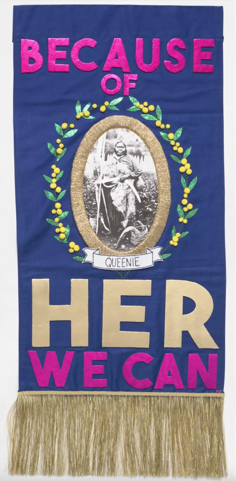 Purple embroidered tapestry containing the words 'Because of her, we can', complete with a photo of First Nations woman Queenie. Purple embroidered tapestry containing the words 'Because of her, we can', complete with a photo of First Nations woman Queenie.