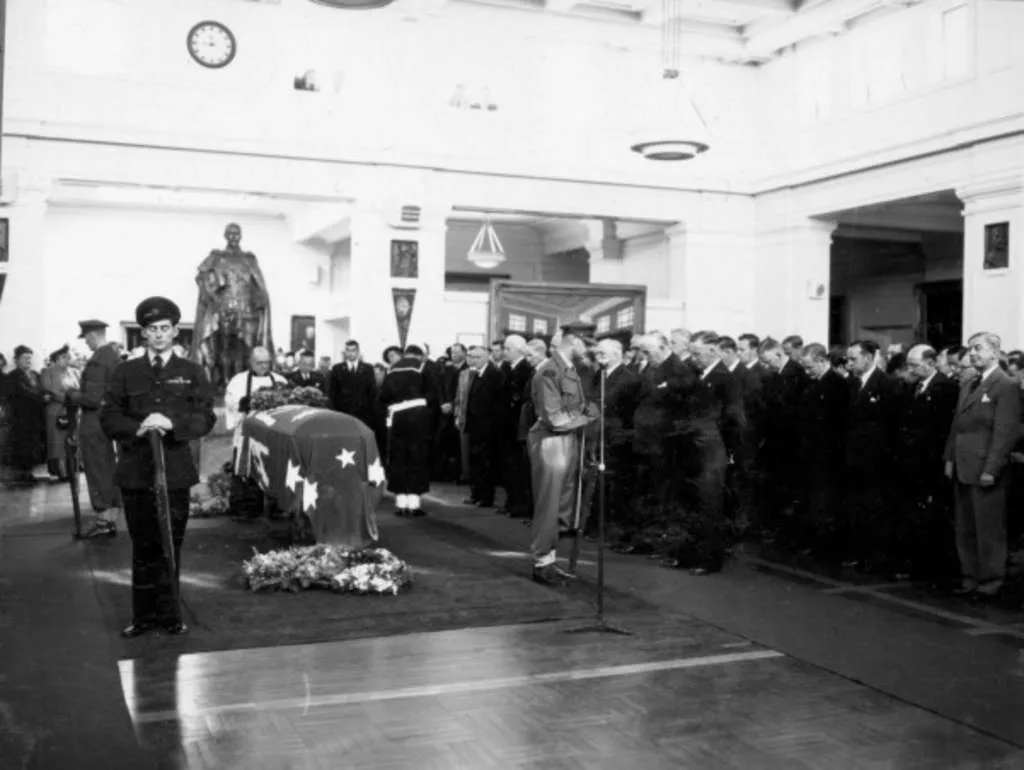 Four armed servicemen stand around a coffin draped in the Australian Flag, whilst a large group of men stand in rows, their heads bowed. Four armed servicemen stand around a coffin draped in the Australian Flag, whilst a large group of men stand in rows, their heads bowed.