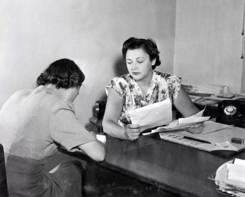 Nancy Wake, a short-haired woman, sitting at a desk, holding papers in her hand. Nancy Wake, a short-haired woman, sitting at a desk, holding papers in her hand.